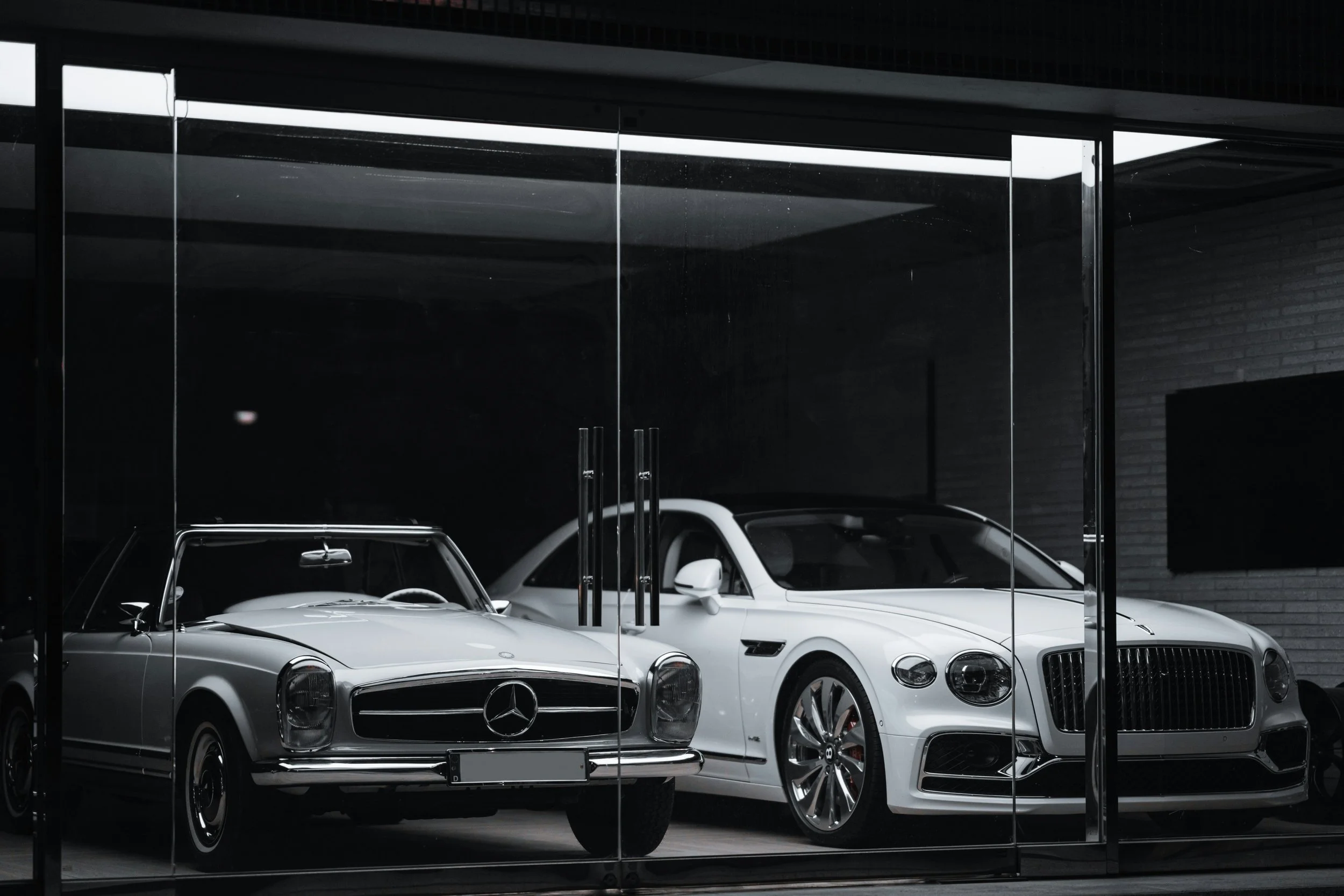 Two classic cars, a silver Mercedes-Benz and a white Chevrolet Corvette, parked inside a showroom or garage, viewed through glass with reflections.