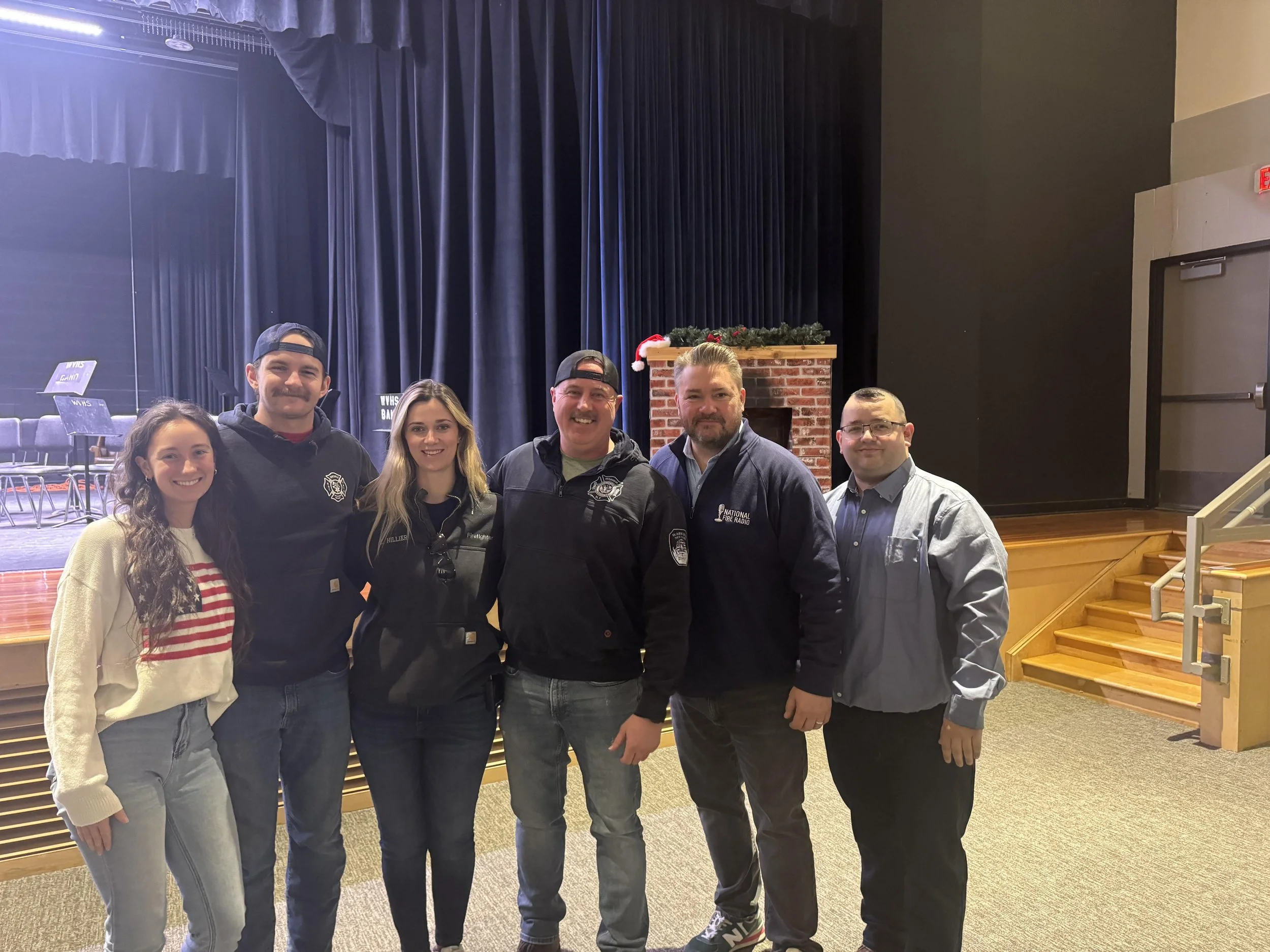 Group of six people standing together on stage, smiling, in a theater or auditorium with blue curtains and a decorated fireplace in the background.