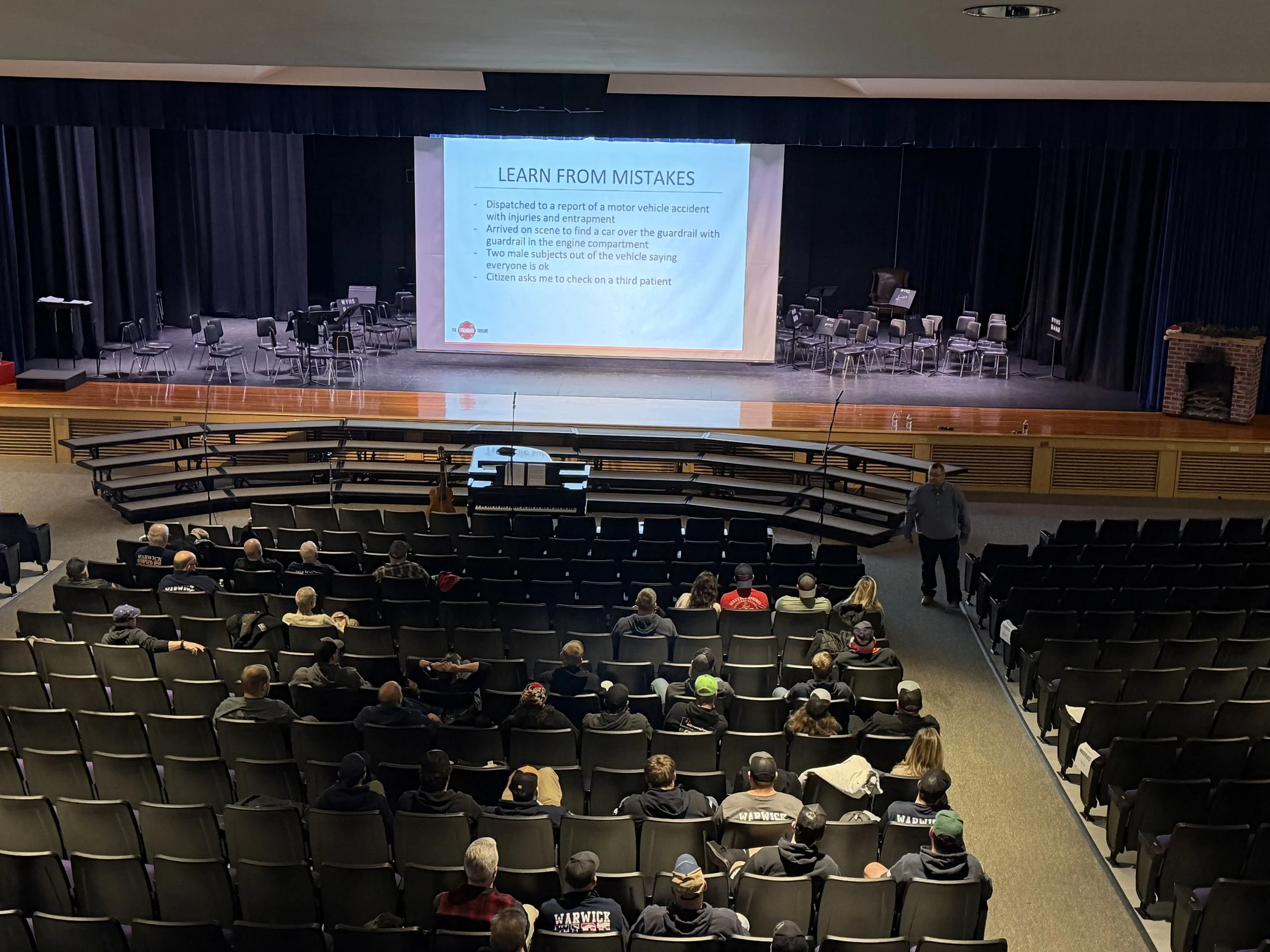 A man gives a presentation to an audience in a theater, with a large screen displaying a slide titled "Learn from Mistakes". The slide lists points about emergency response and accident management. The theater is partially filled with audience members seated facing the stage, with chairs and musical instruments on the stage.
