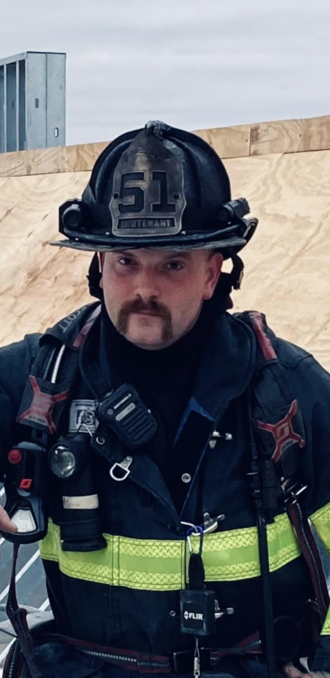 Firefighter in uniform with a helmet marked '51 LUTHERAN' and a radio, standing in front of a wooden construction frame.
