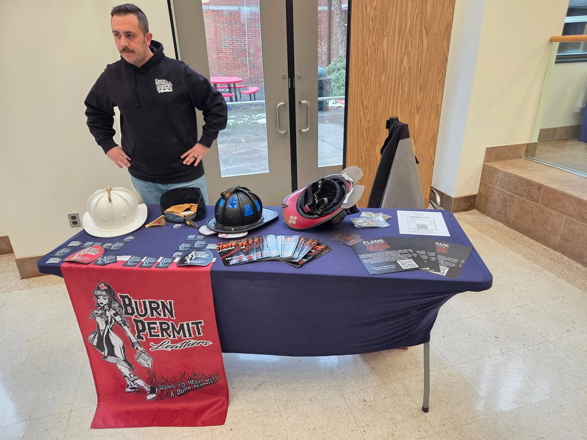 Man standing behind a table with promotional materials for Burn Permit Leathers, featuring motorcycle helmets, flyers, and a flag. The table is inside a building near glass doors and a staircase.
