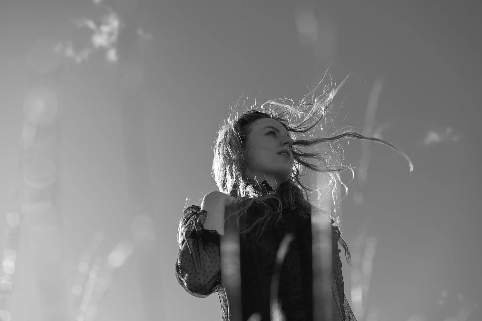 A black and white photo of a woman with windblown hair looking up, with a partly cloudy sky in the background.
