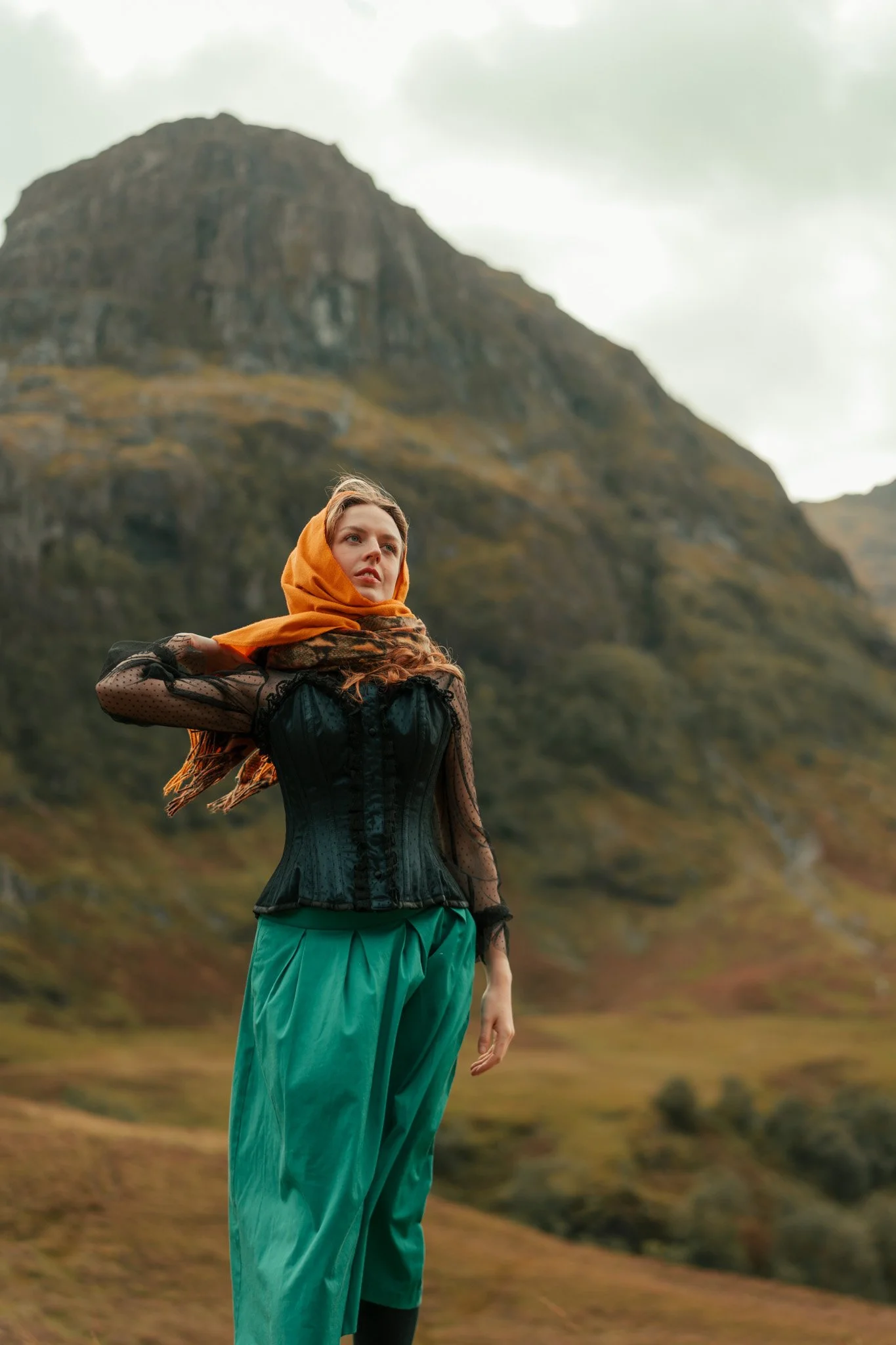 A woman with long hair standing outdoors in front of a mountain landscape, wearing a black lace top, orange scarf, and teal pants.