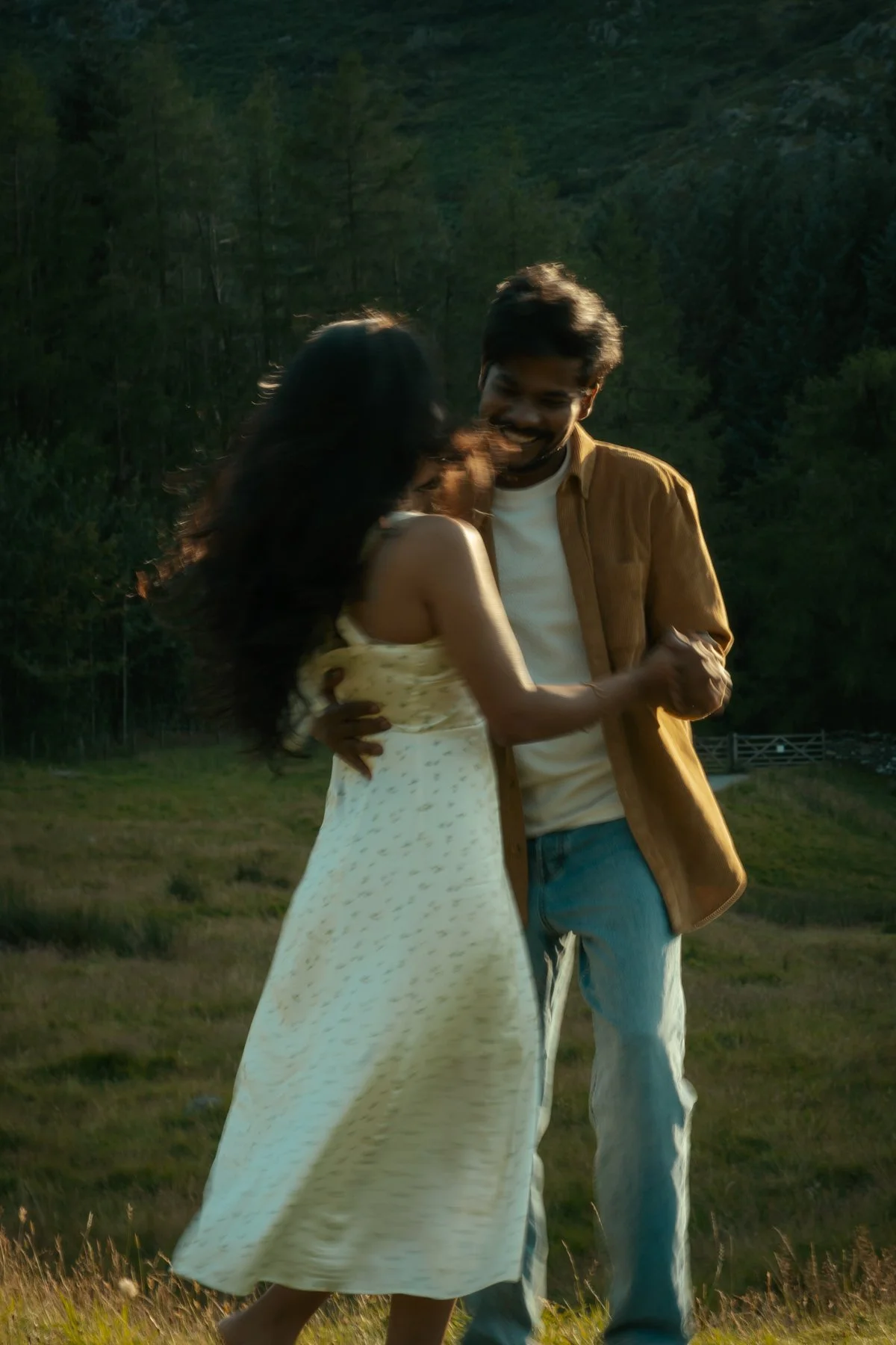 A man and woman dancing outdoors on a grassy field during sunset, with a forested mountain in the background, smiling and enjoying each other's company.