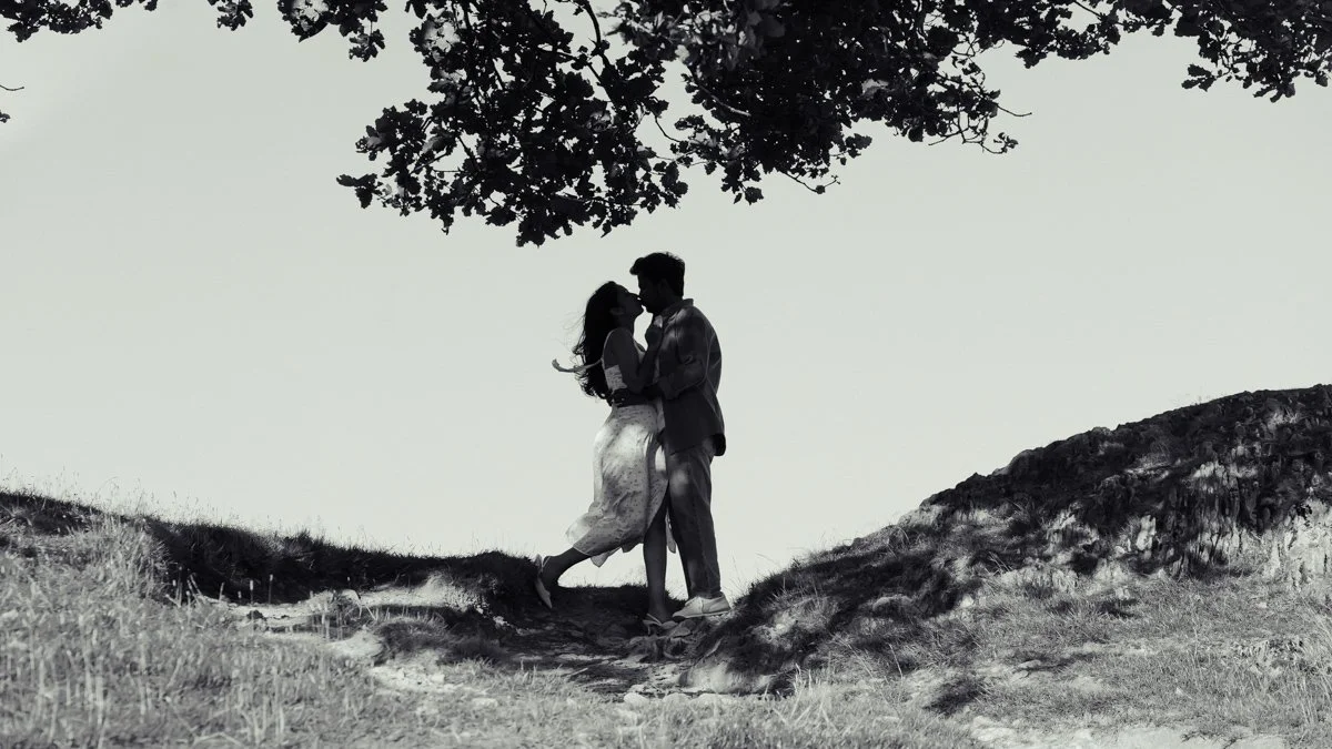 A man and woman in wedding attire sharing a kiss in a forest with a waterfall in the background.