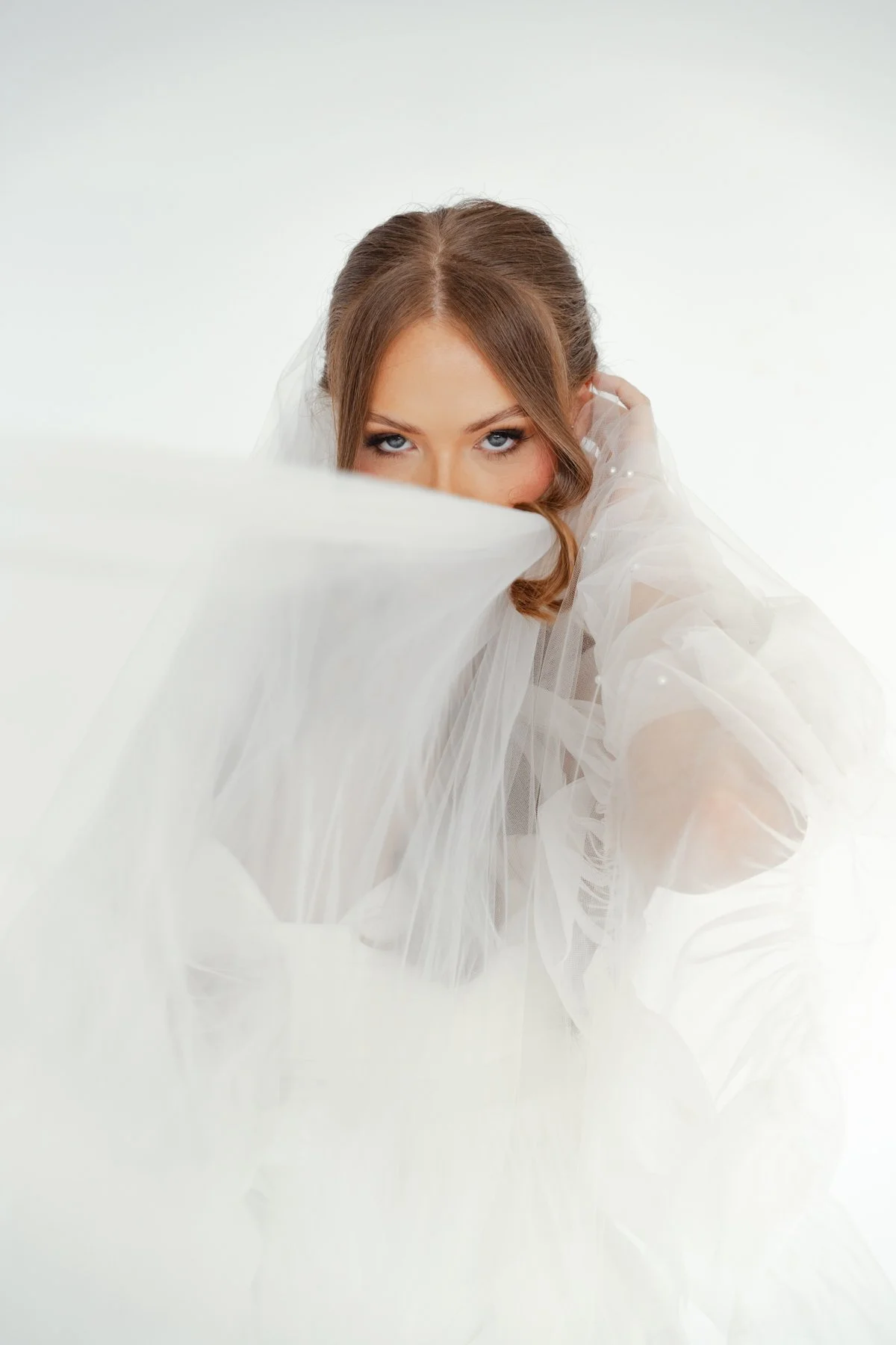 A woman with brown hair, styled in waves, gazes over a white veil she is holding, with piercing blue eyes and a soft expression, against a plain white background.
