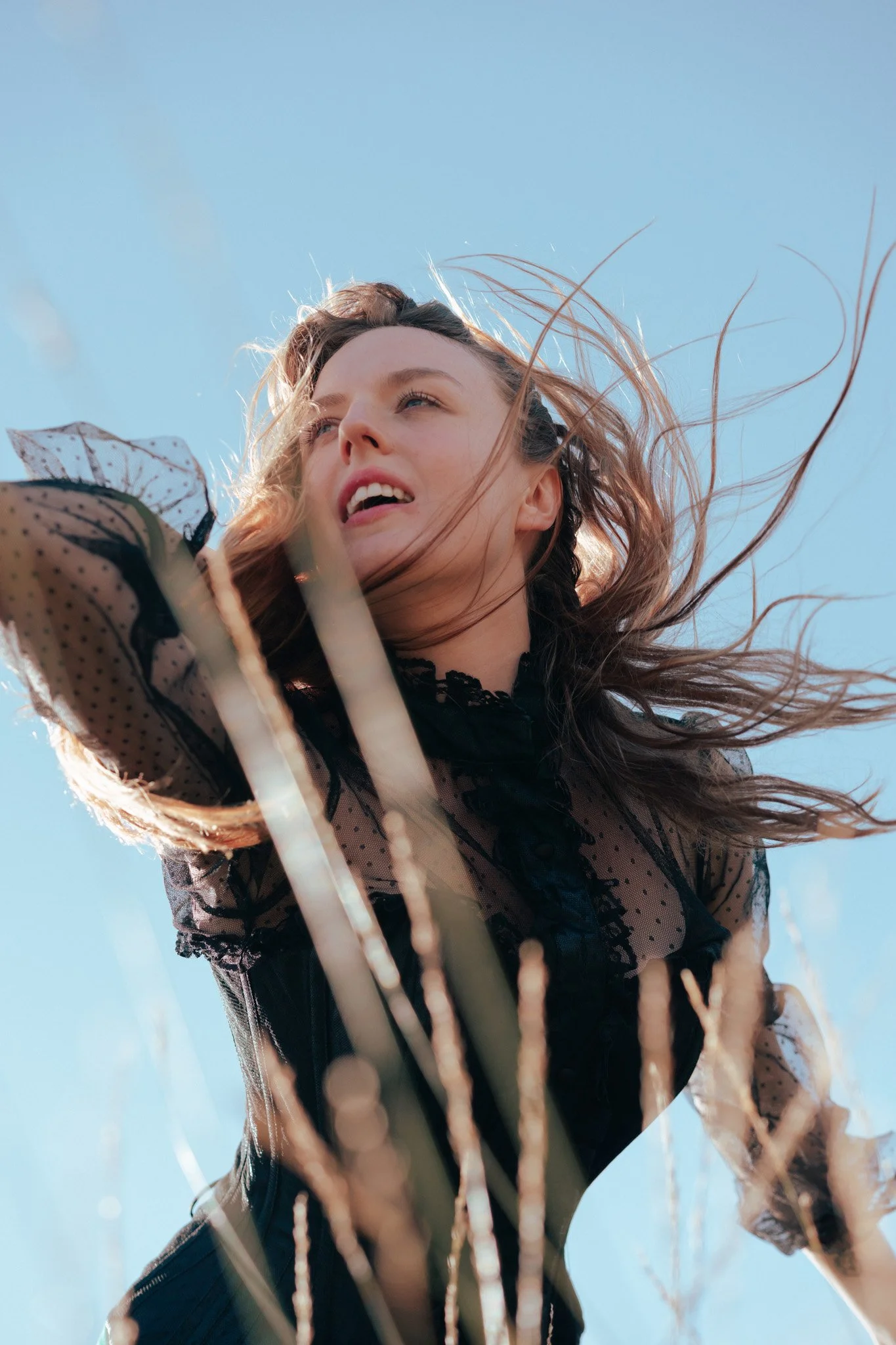 A young woman with wavy hair outdoors, looking upward with a blue sky in the background, wearing a black lace top.