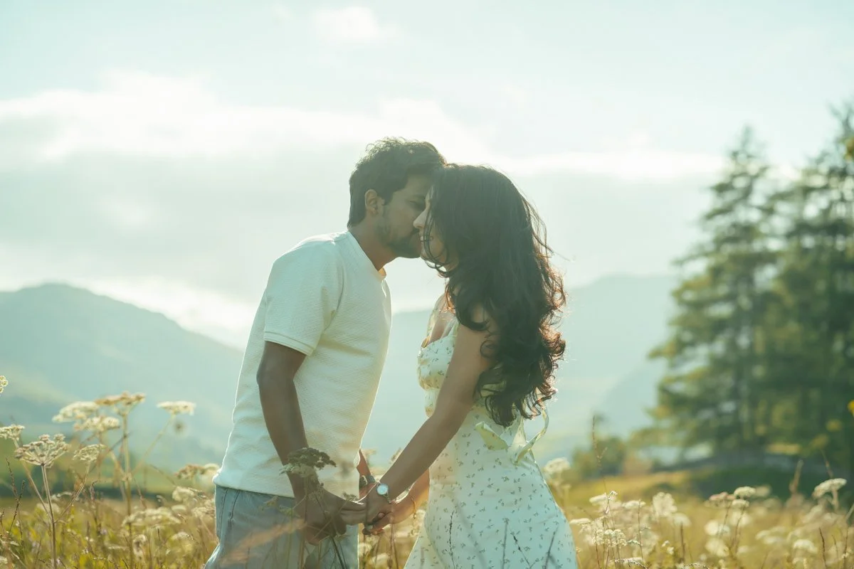 A couple stands close together in a field of tall flowers, holding hands, with their foreheads touching, against a backdrop of mountains and a cloudy sky.