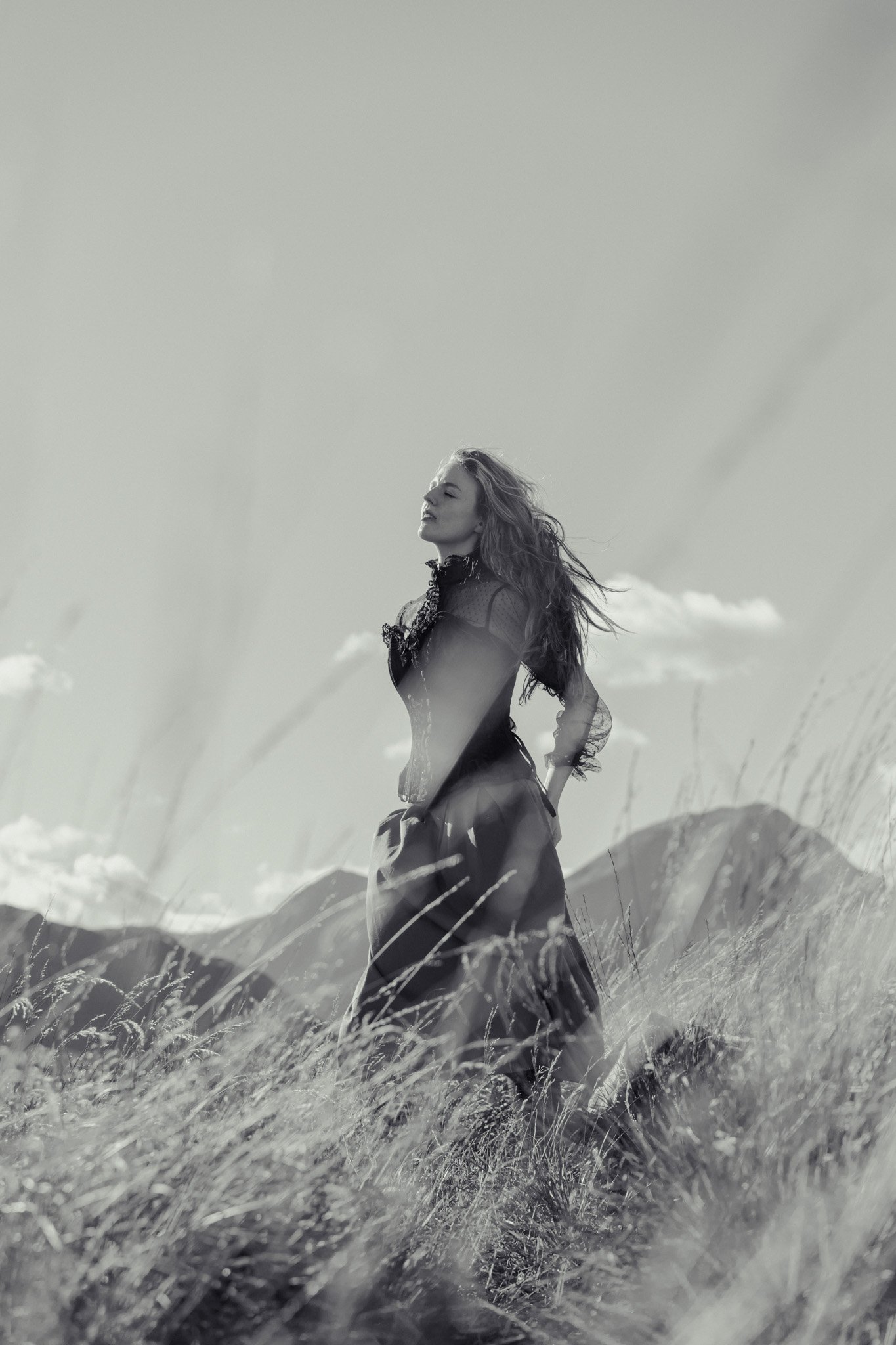 A woman in a long, flowing dress standing in a grassy field with mountains and a cloudy sky in the background in black and white.