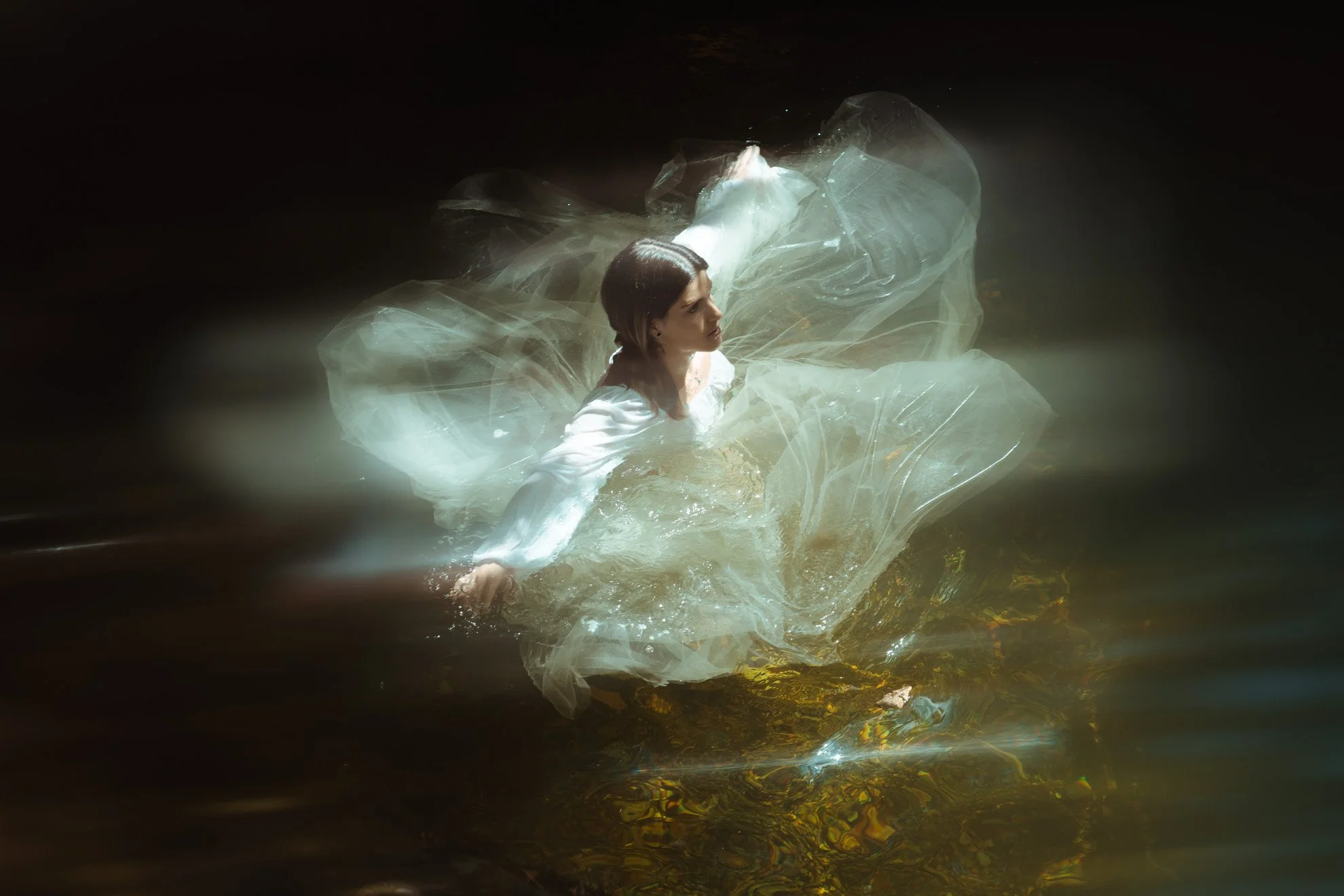 A woman in a white dress is submerged in water and appears to be floating or swimming, with dark surroundings.
