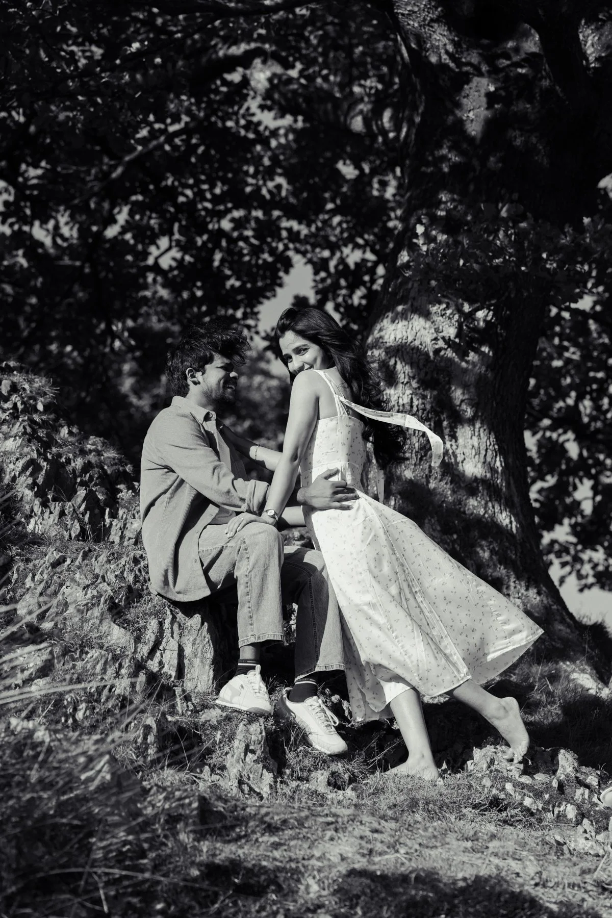 A black-and-white photo of a couple sitting and smiling on a rock outdoors, surrounded by trees and foliage.