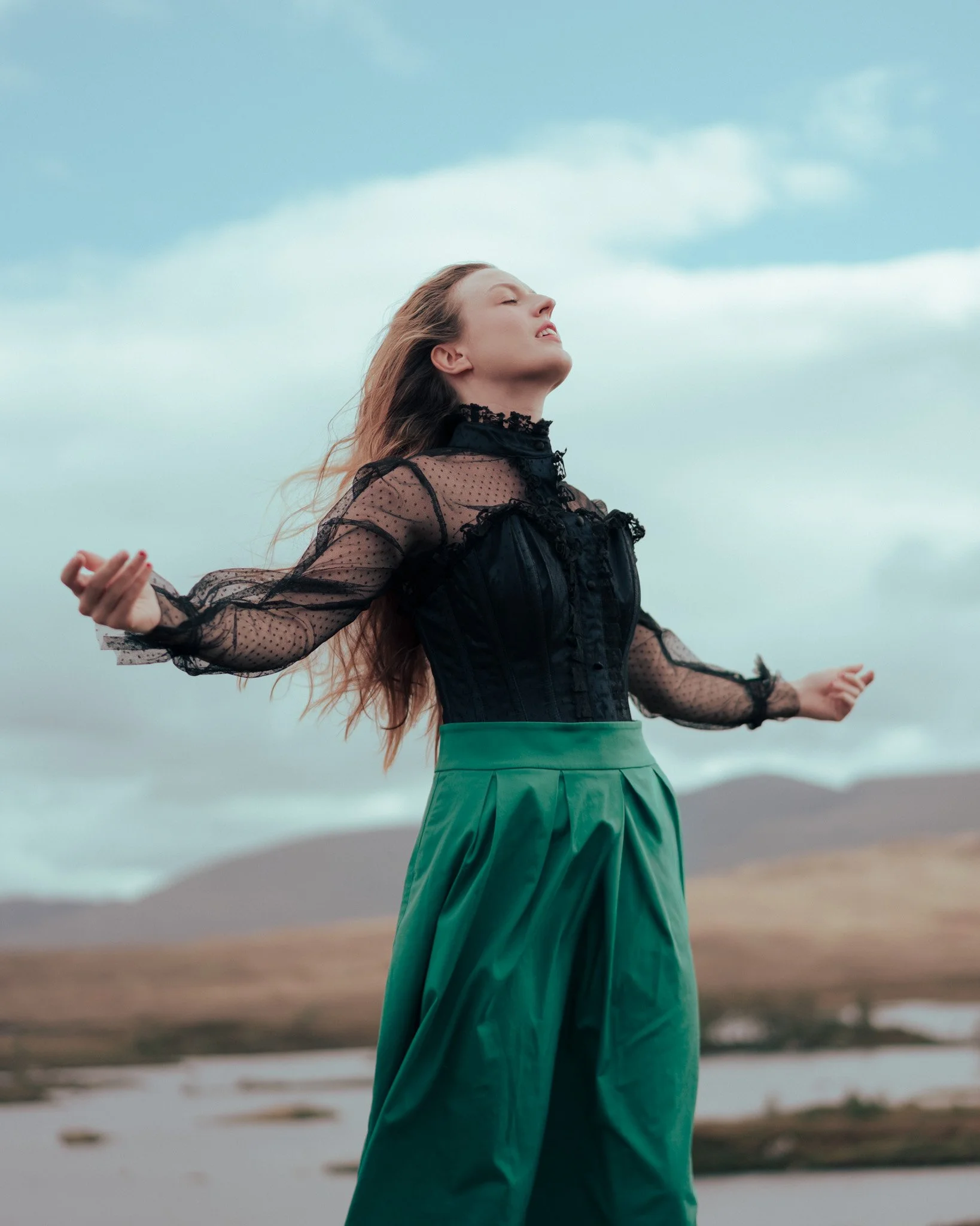 A woman with closed eyes and open arms standing outdoors with a cloudy sky and mountains in the background