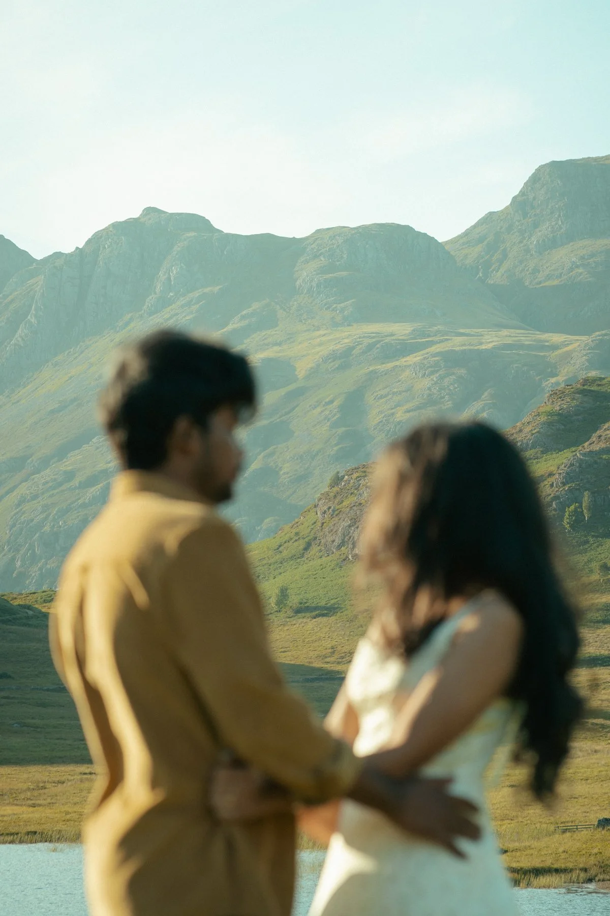 A man and woman standing close together outdoors in front of green mountains, holding hands, with both slightly blurred.