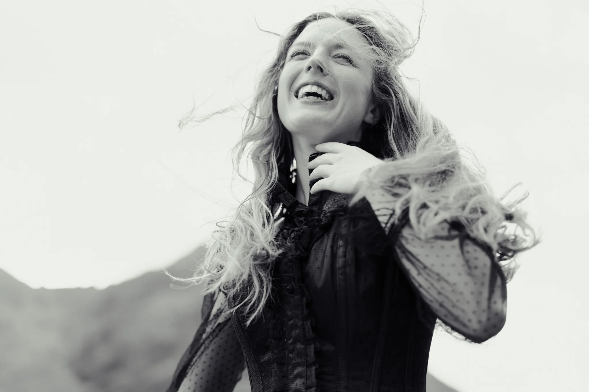 A woman with long, curly hair is laughing outdoors, holding her hand near her neck, laughing joyfully