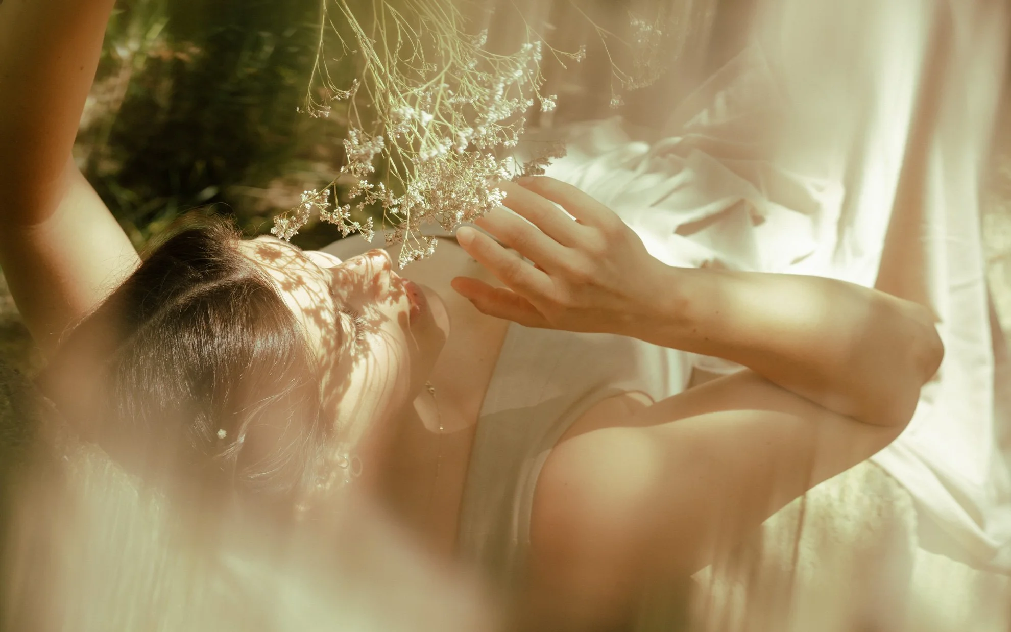A  dreamy portrait of a woman lying on her back outdoors in sunlight, with her arm raised above her head and her hand resting on her forehead, holding a bunch of dried flowers.