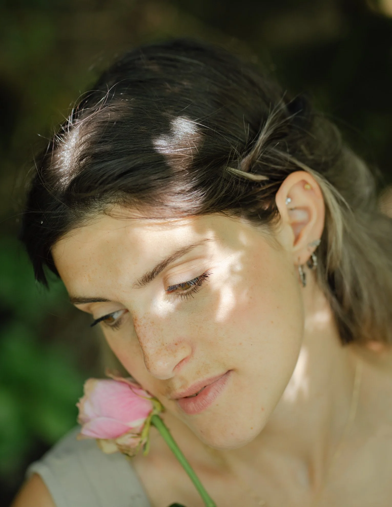Close-up of a woman with dark hair and light skin, gently resting her head near a pink flower
