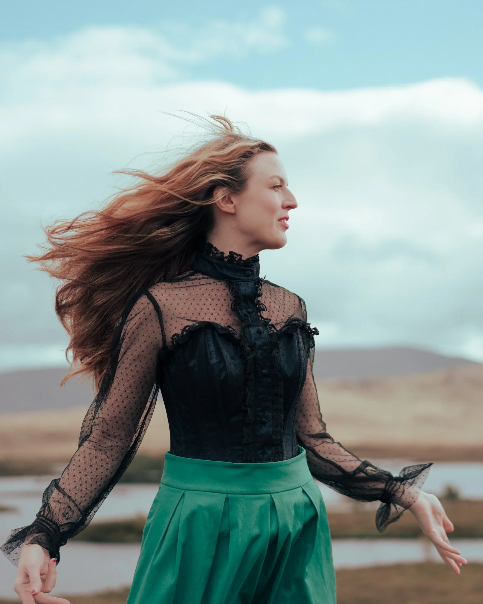 A woman with long hair flowing in the wind, wearing a sheer corset and high-waisted green skirt, windswept by the mountain winds