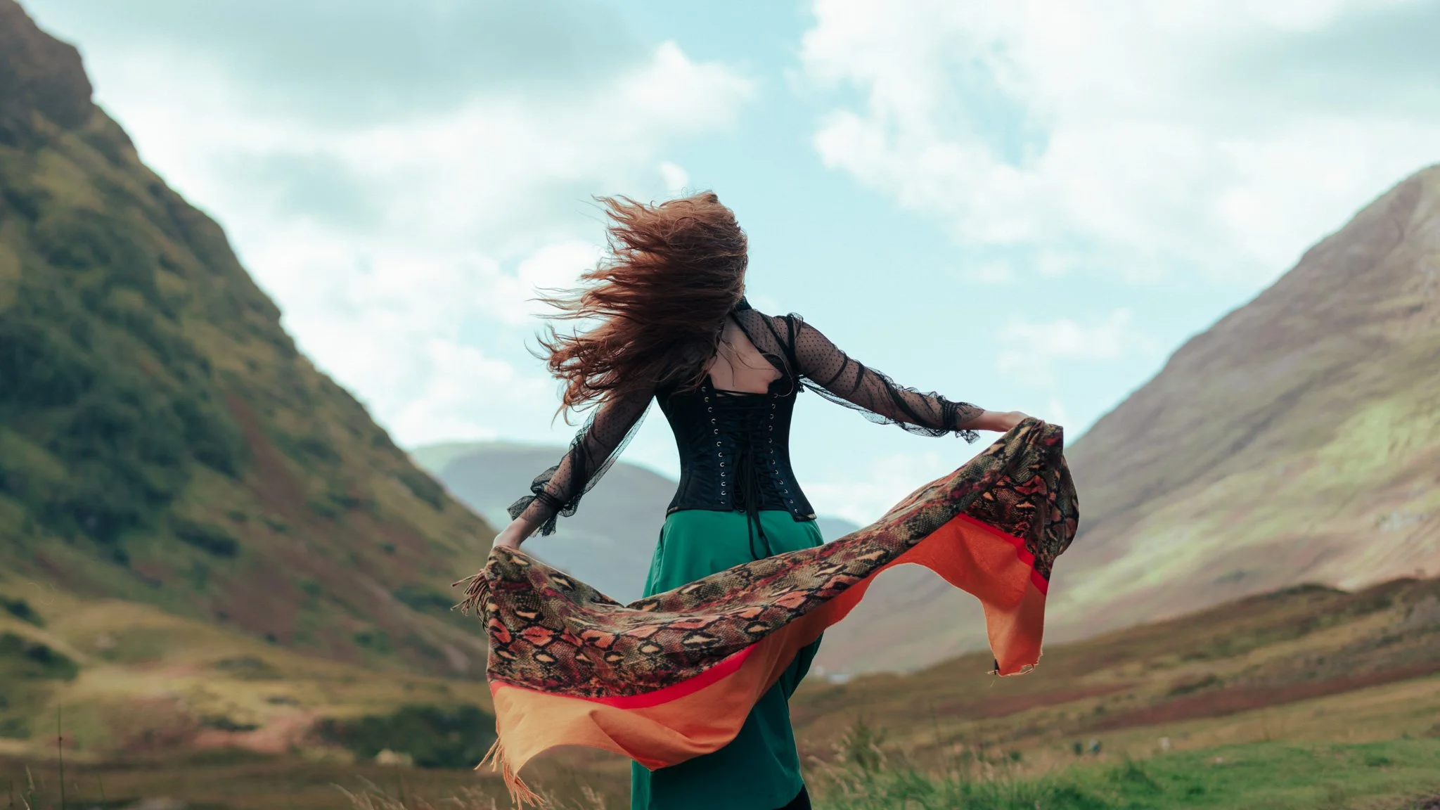 A woman standing in a green valley with mountains in the background, holding a colorful scarf and her hair blowing in the wind.