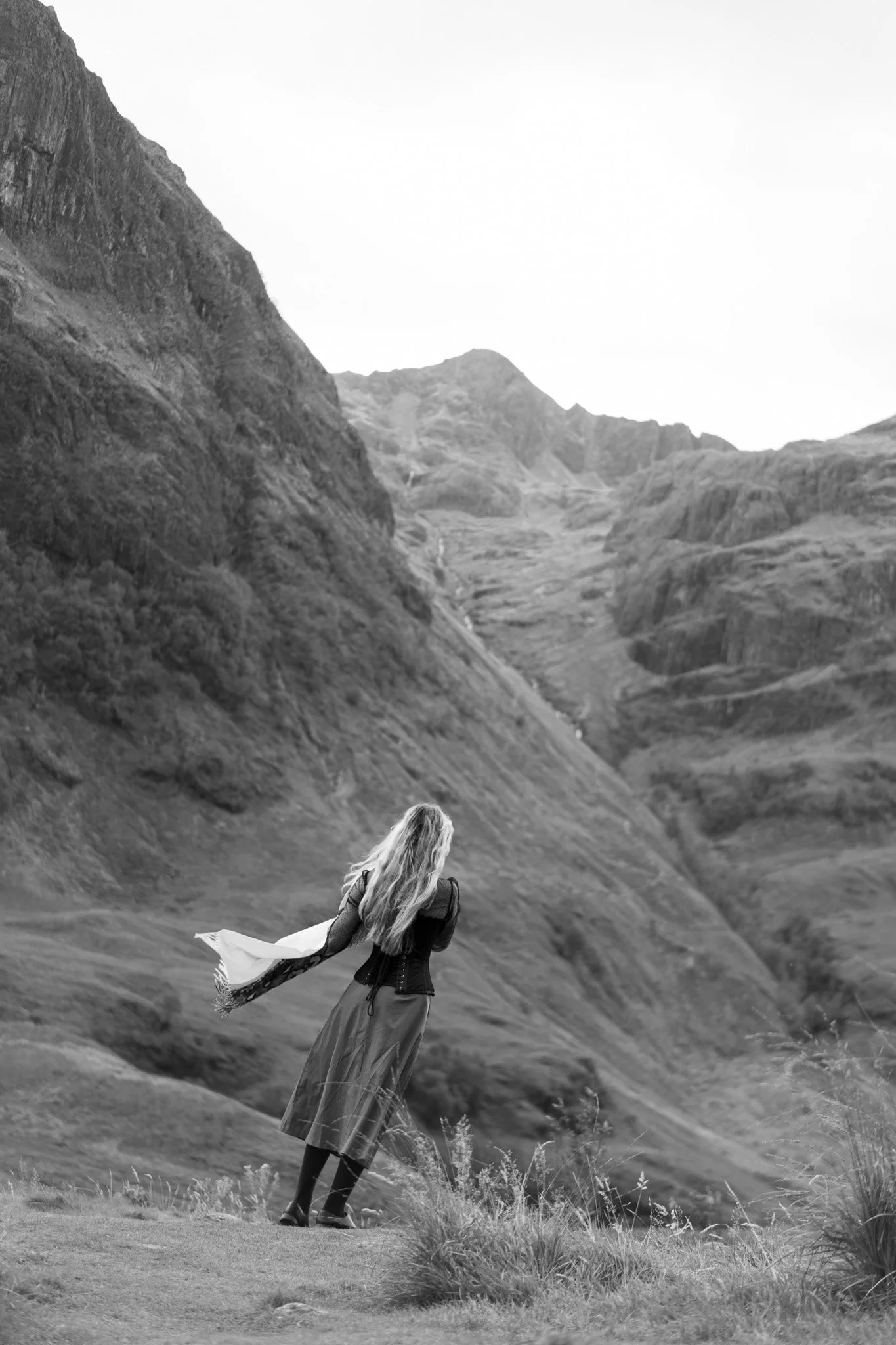 A woman with long hair wearing a long dress and a coat is suspended in free fall at the edge of the mountain
