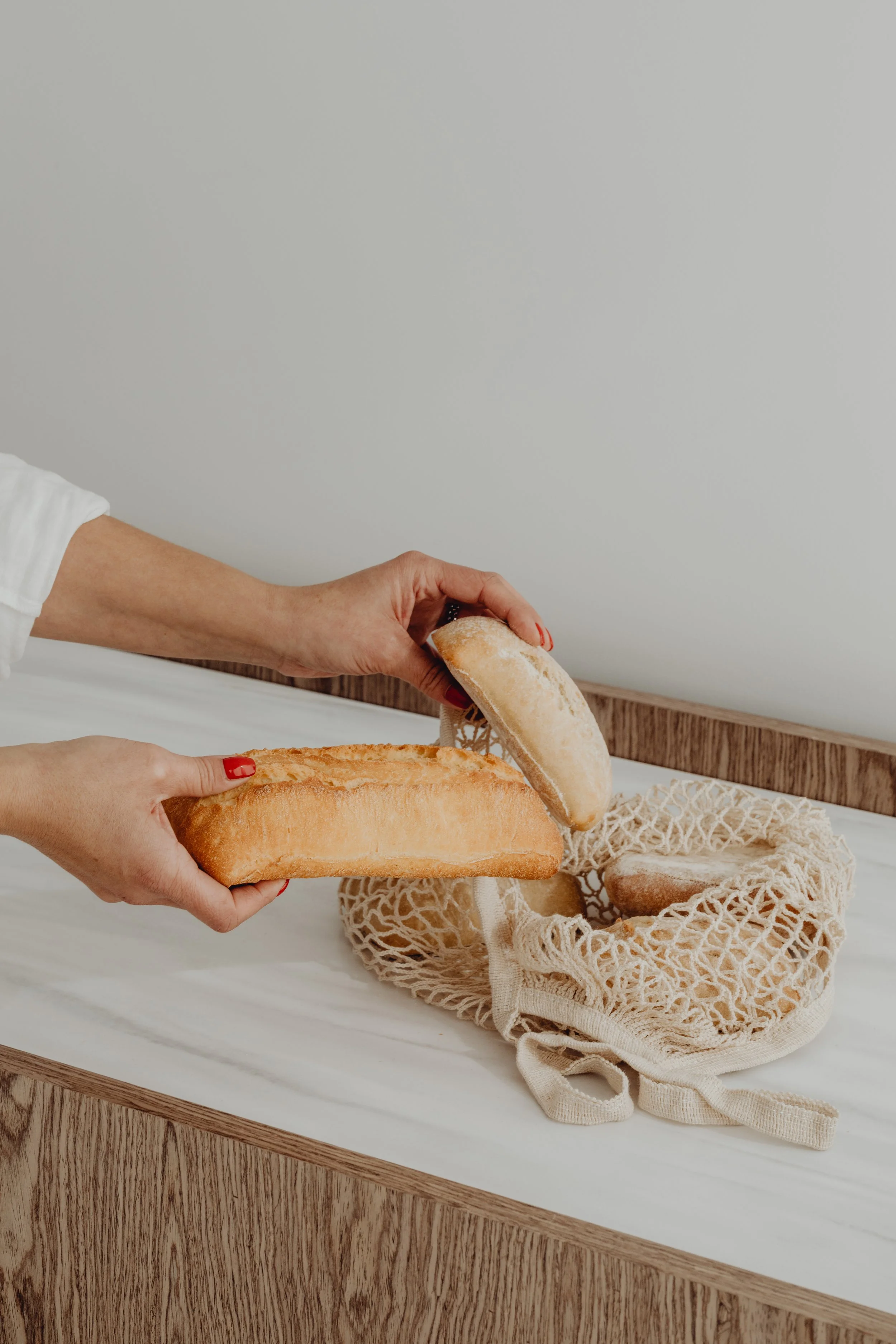 Hands holding a baguette and a piece of bread over a reusable cotton mesh bag containing other bread loaves on a white surface.
