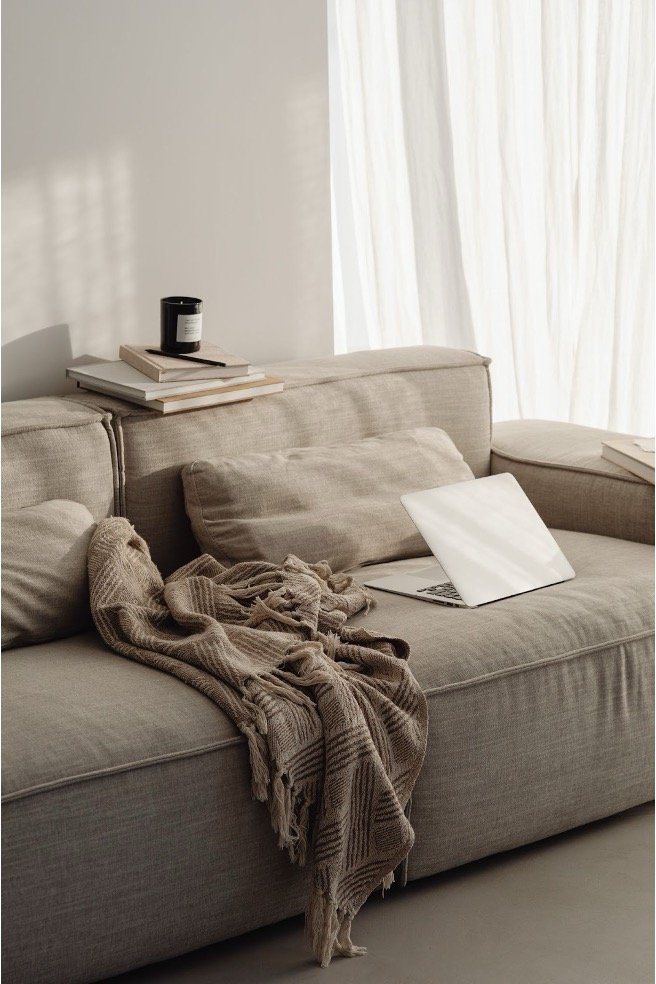 A beige sofa with a blanket, pillows, a laptop, and a stack of books on it, next to a window with white curtains.