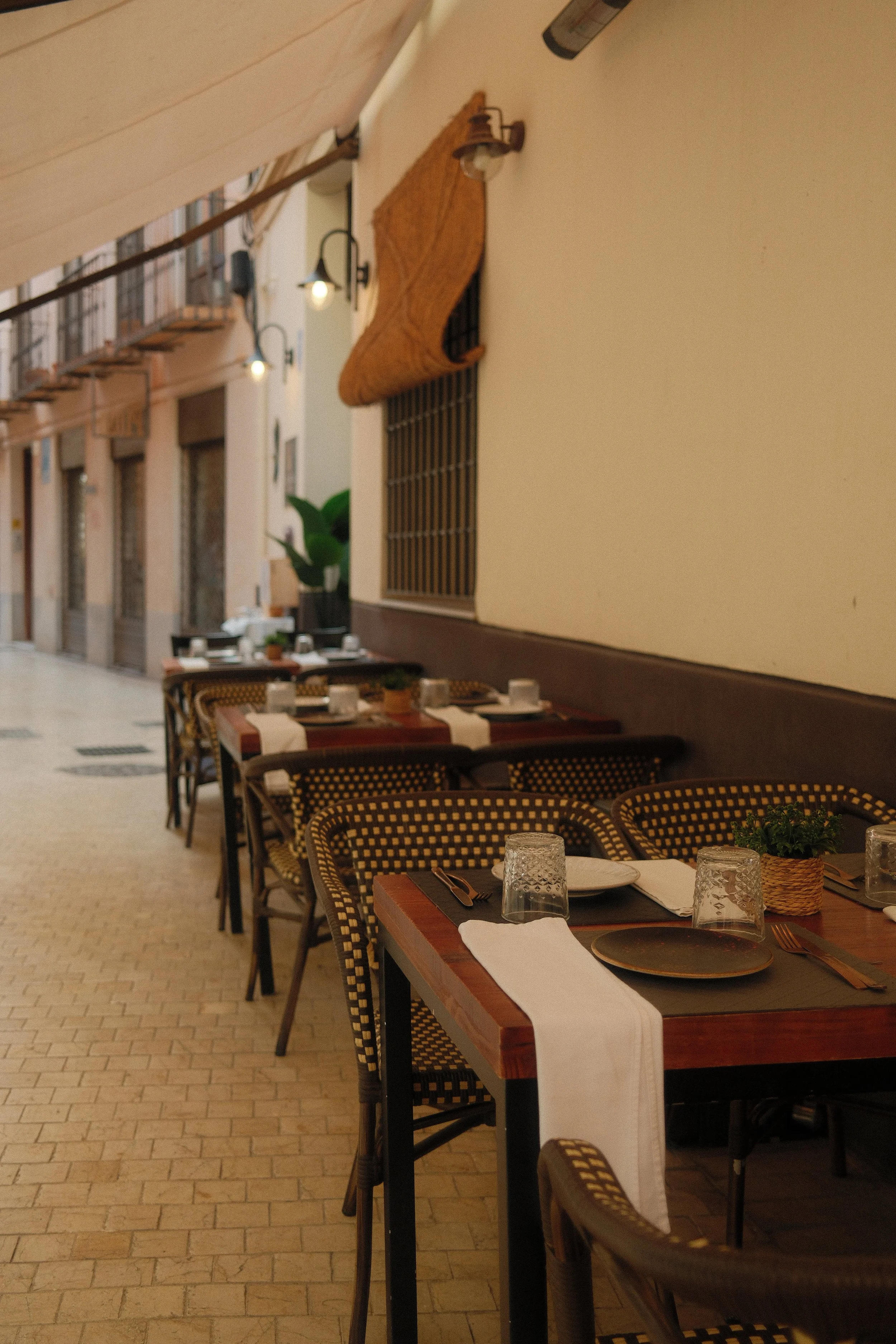 Set dining tables with plates, glasses, utensils, and napkins inside a restaurant with wicker chairs, potted plants, and wall decorations.