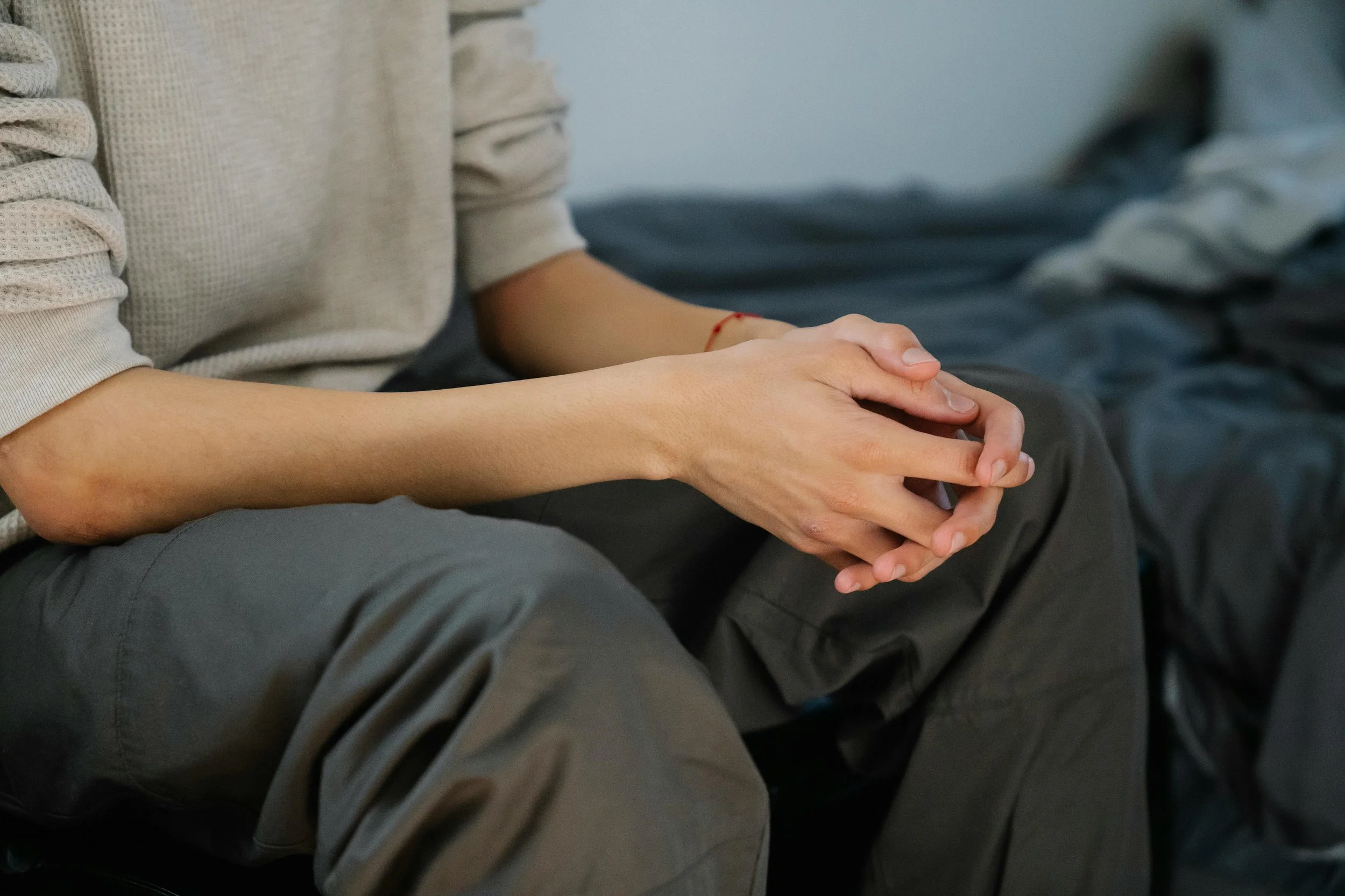 A person sitting on a bed, wearing a beige long-sleeve shirt and dark gray pants, with clasped hands resting on their lap.
