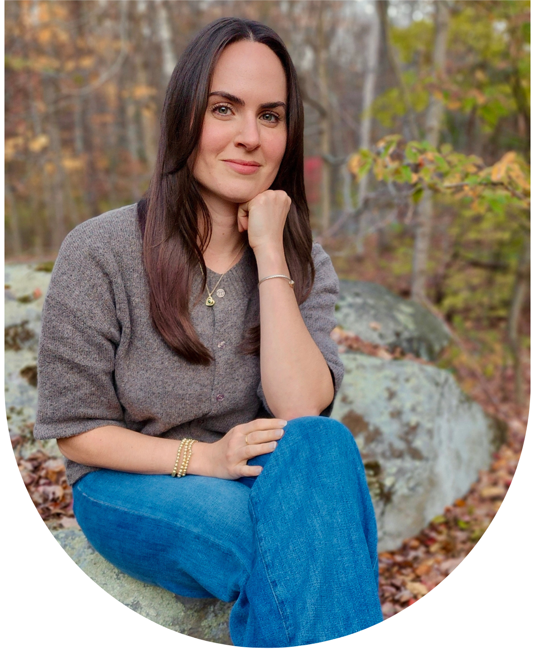 A woman with long brown hair sitting on a rock in a wooded area during autumn, wearing a gray blouse and blue jeans, with her chin resting on her hand, smiling softly.