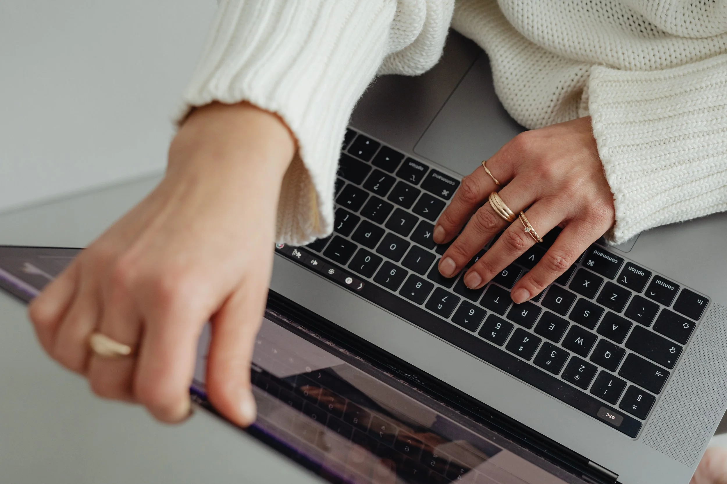 Person in a cream sweater using a laptop, with hands on keyboard and touchscreen, wearing multiple rings.