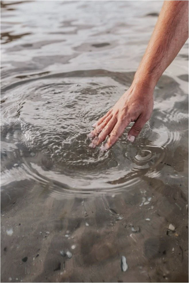A person's hand is touching water in a natural setting, creating ripples on the water surface.