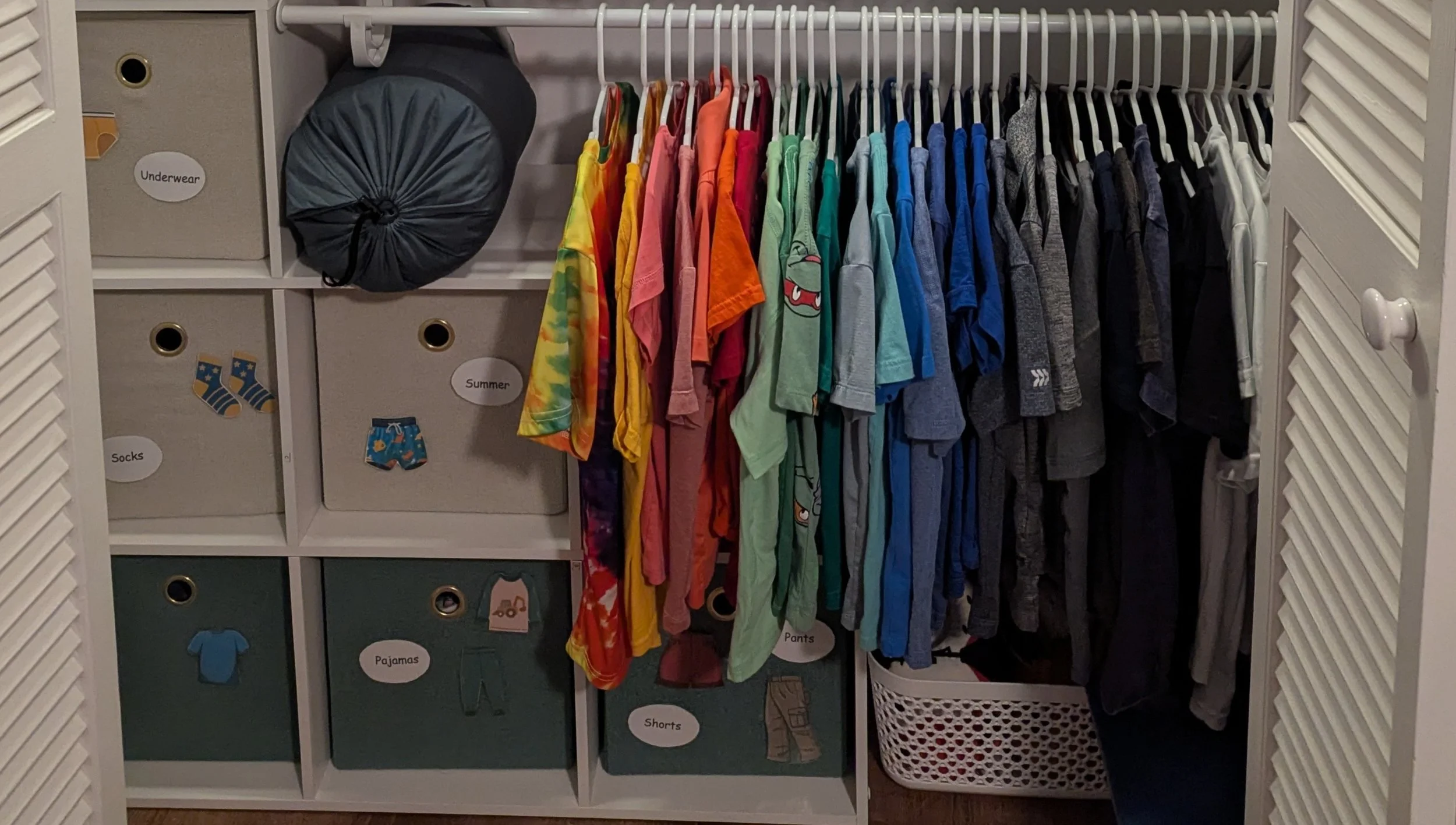 Inside a closet with organized shelves labeled for underwear, socks, pajamas, shorts, and summer clothing. Hanging on a rack are children's colorful t-shirts and pajamas, with a laundry basket at the bottom.