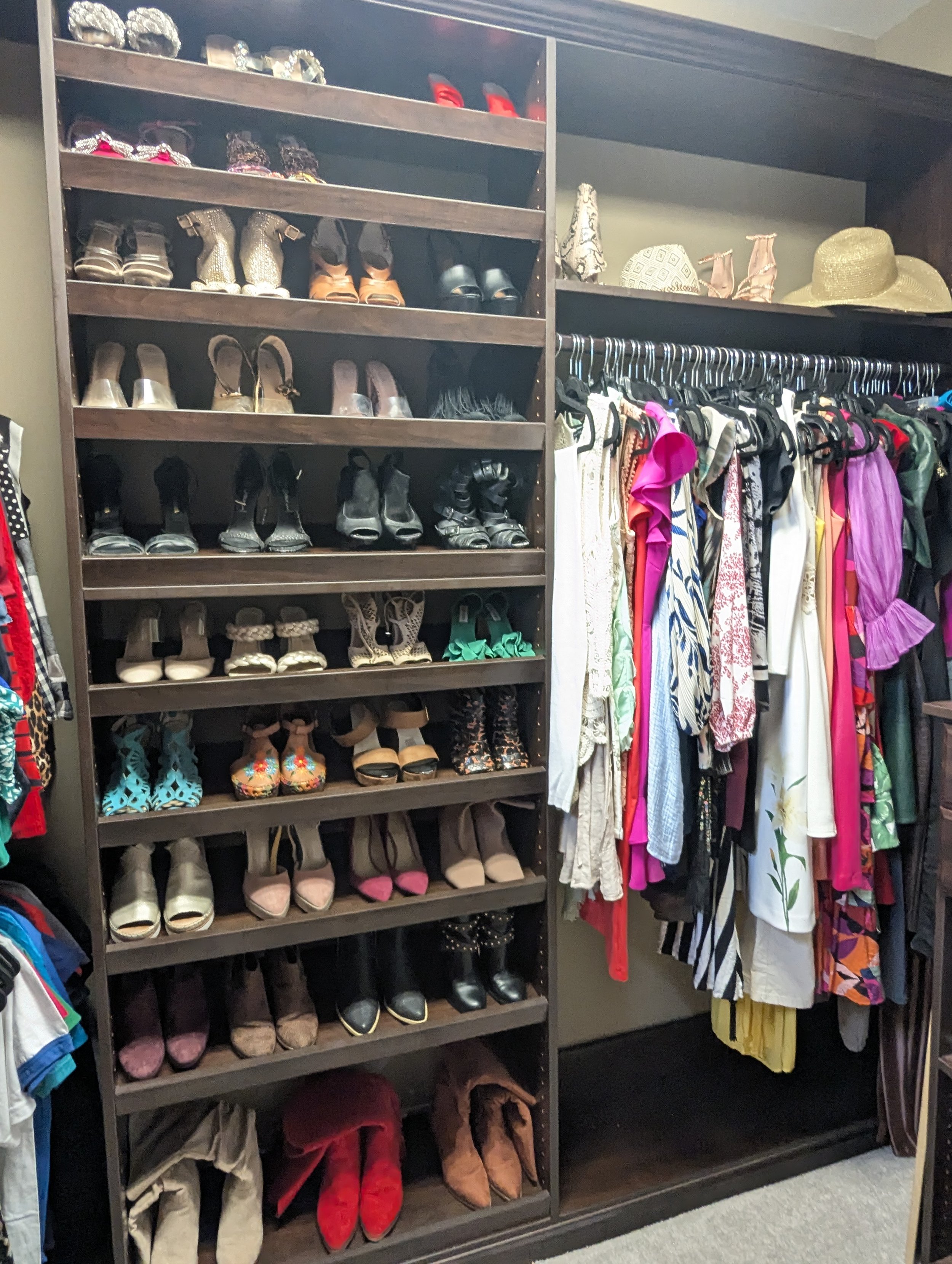 Closet with multiple shelves filled with various women's shoes, including heels, sandals, and boots, next to a hanging rod with colorful dresses, skirts, and tops, along with some hats on top.