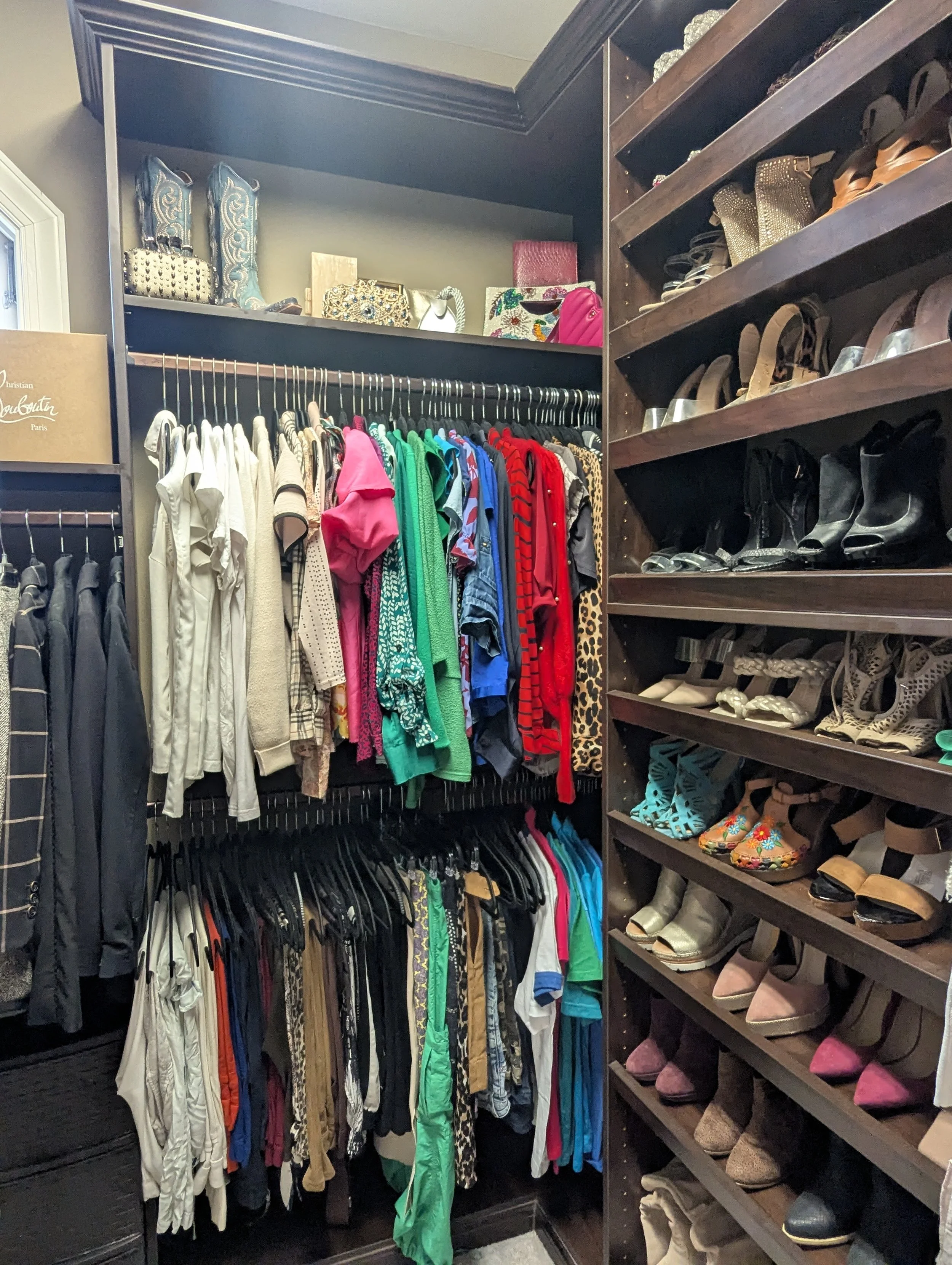 Closet with clothing and shoes. Top shelf has handbags and boots. Middle section has hanging clothes including shirts, dresses, and jackets in various colors. The bottom has more clothes on hangers. To the right, multiple shelves hold high heels and 