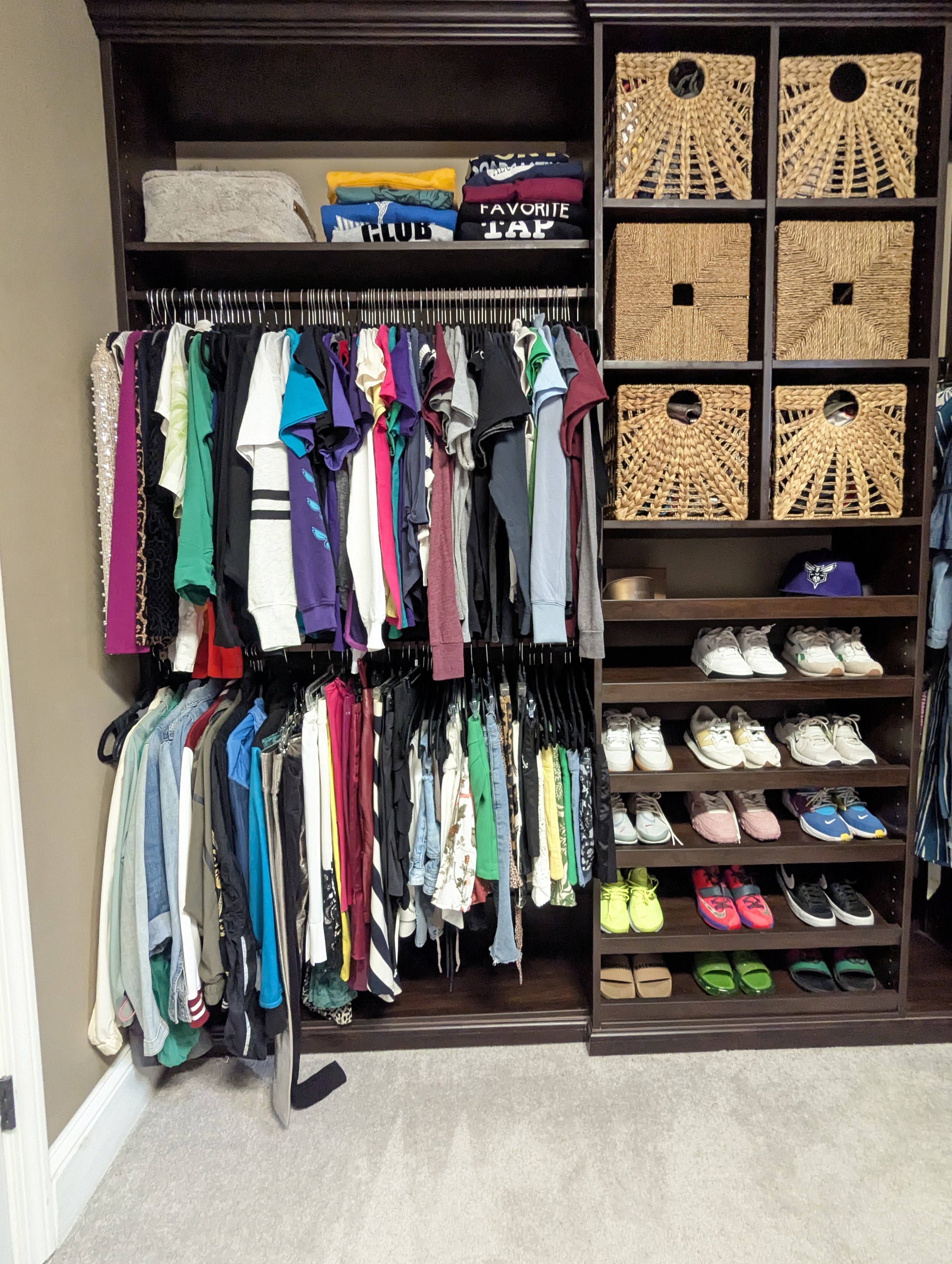 A closet with a shelf of folded clothes, a hanging rack of various shirts and pants, a shelf with wicker baskets, and a shoe rack with sneakers and sandals.