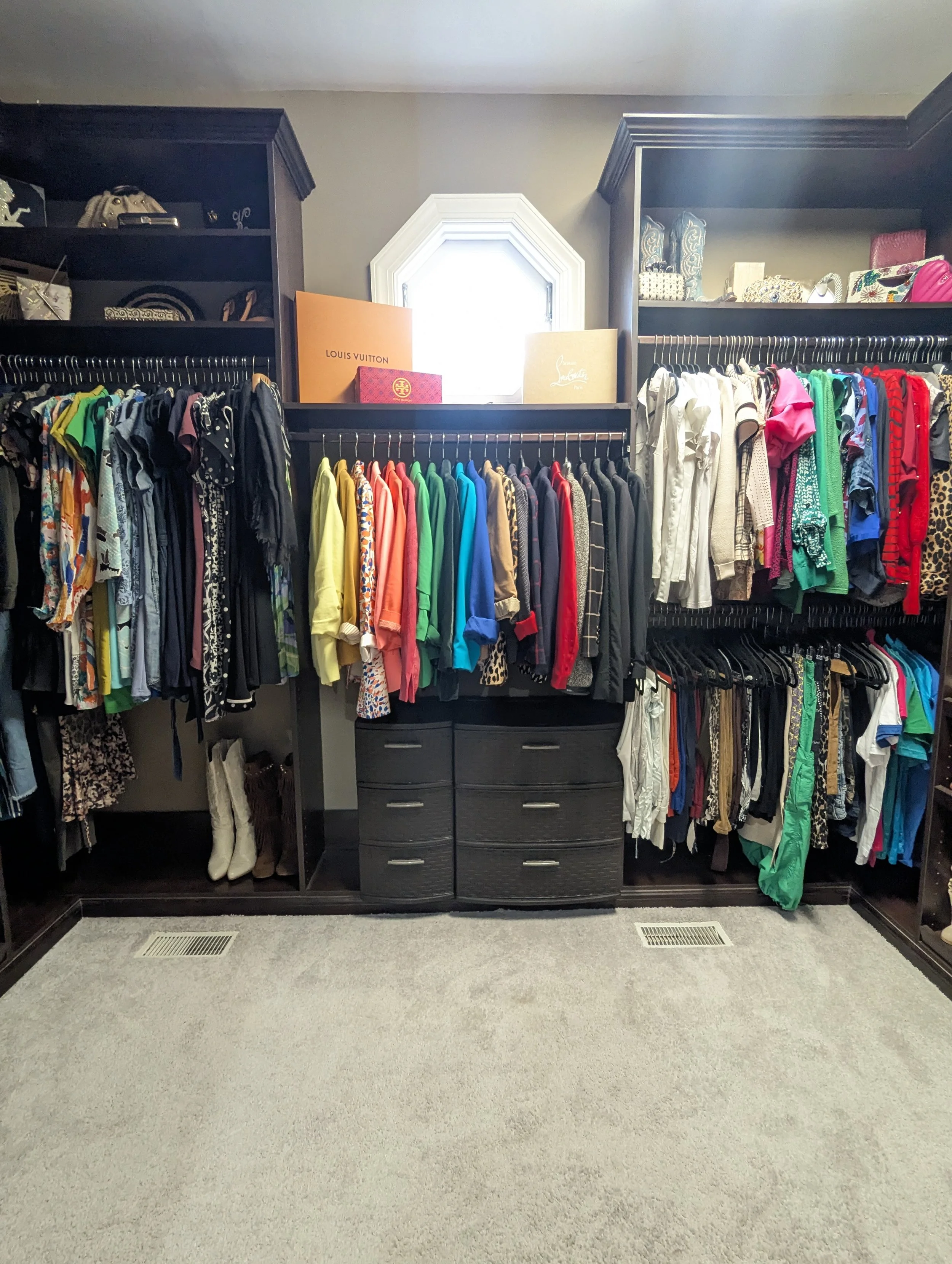 A walk-in closet with racks of colorful clothes, shelves with designer bags and boxes, a small window, and a tan carpeted floor.