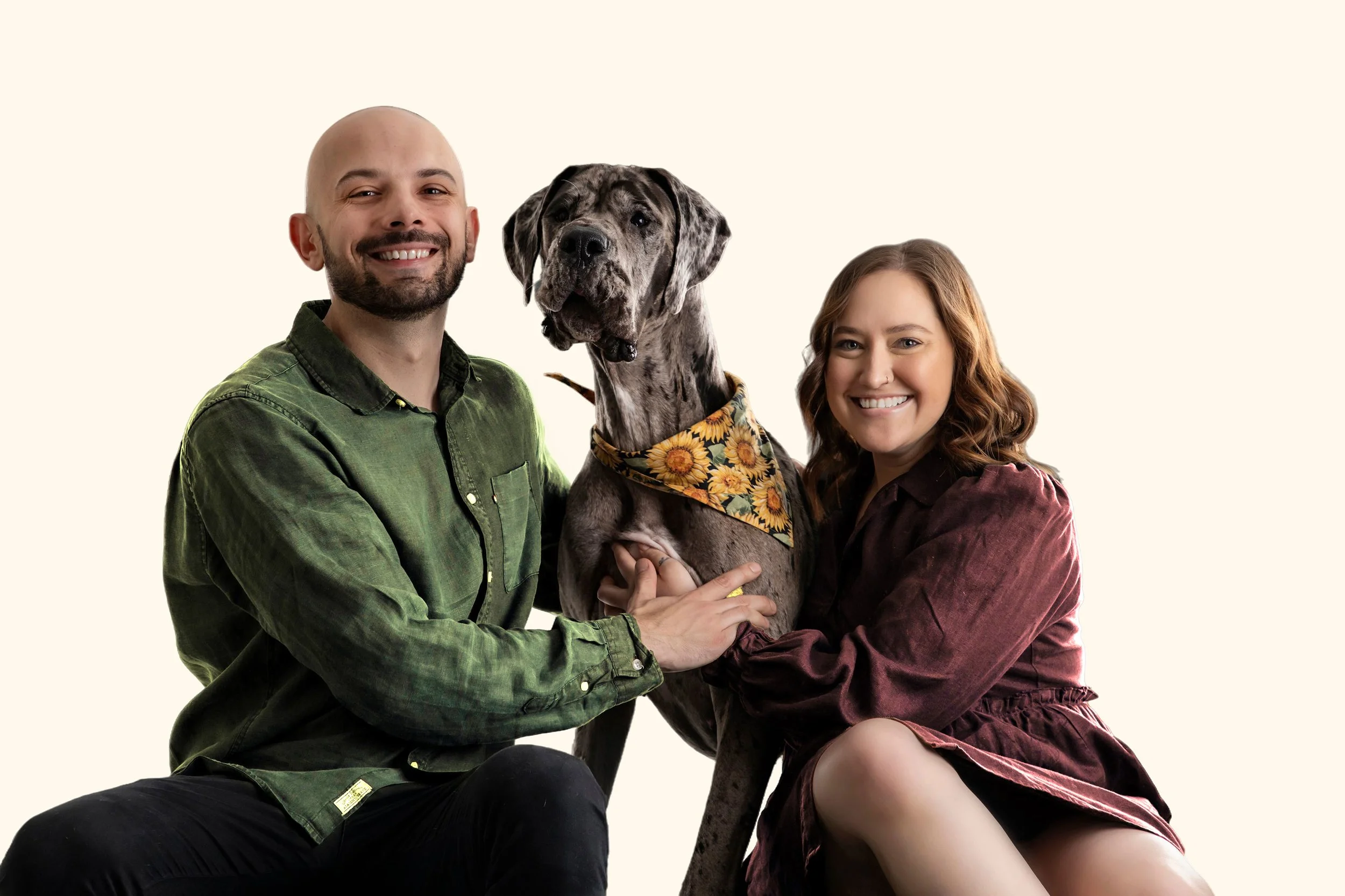 A smiling man and woman posing with their Great Dane dog, which is wearing a sunflower bandana, against a plain light background.