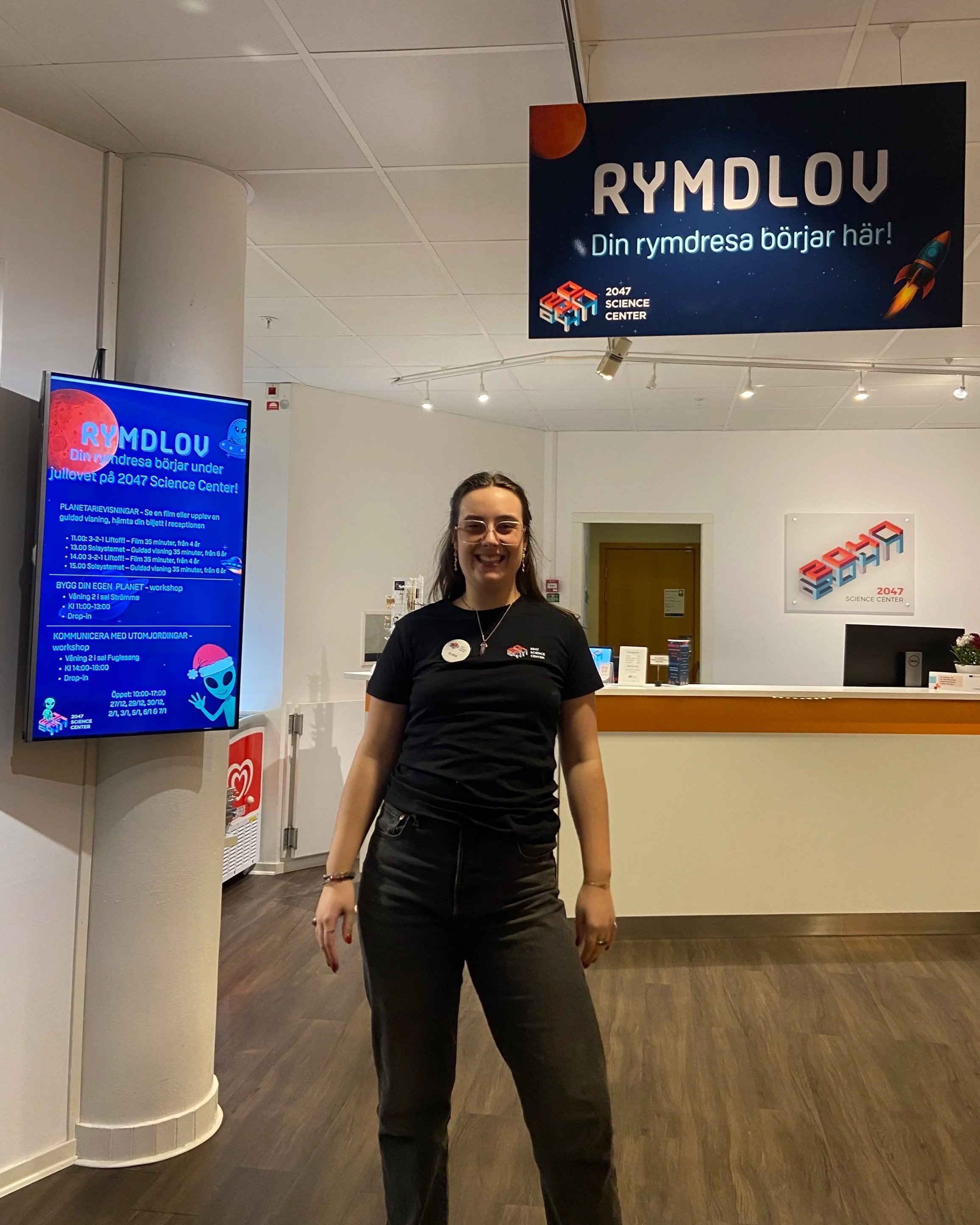 En kvinna med mörkt hår och glasögon står inomhus, framför en reception på en science center. Det finns skyltar ovanför med texten 'RYMDLOV Din rymdresa börjar här!'.
