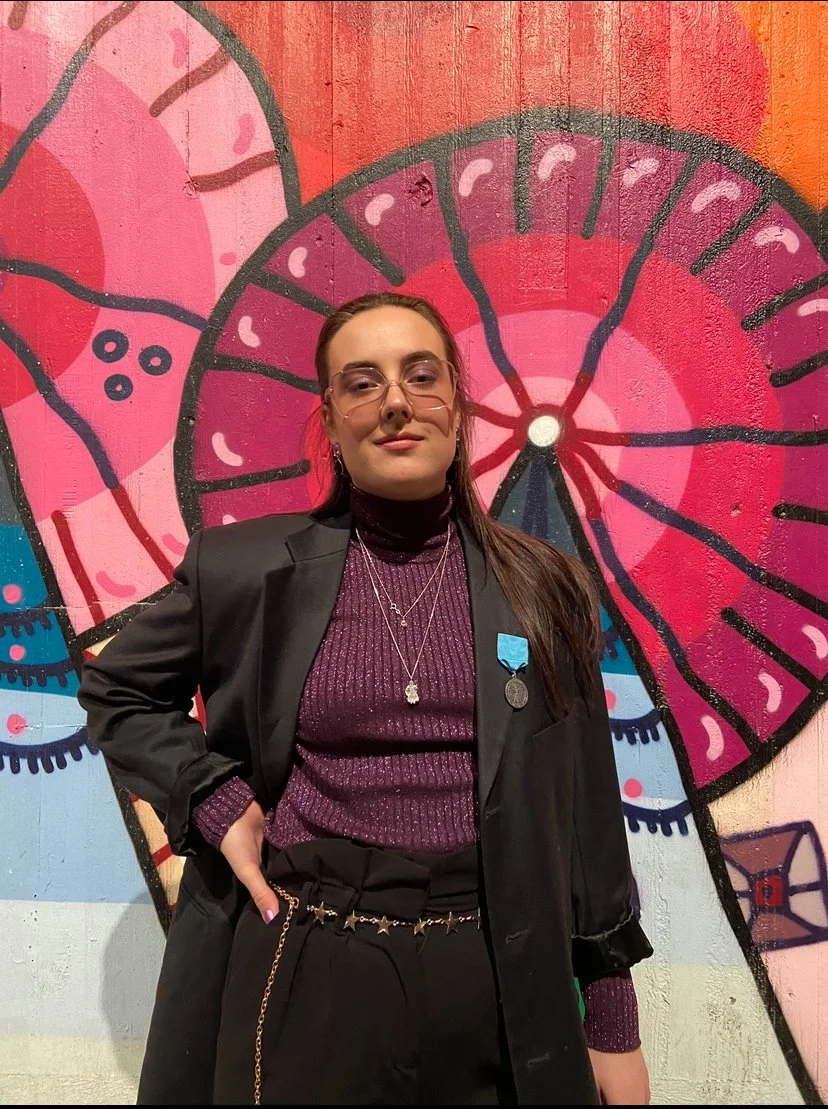 En kvinna står framför en färgglad muralmålning med rosa och blå nyanser. Hon bär glasögon, en svart blazer, en mörk lila turtleneck och har medaljer på kläderna.