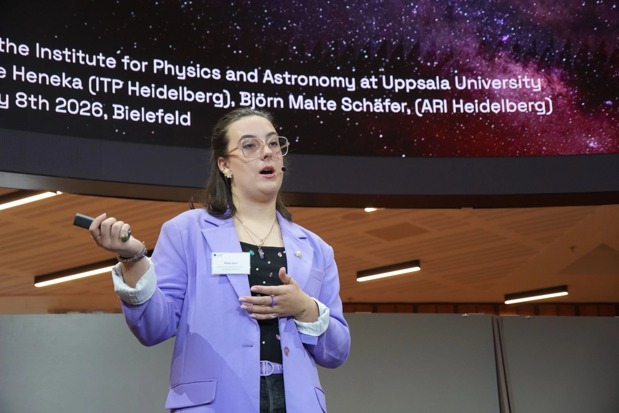 En kvinna i en ljuslila kavaj håller ett mikrofonhandtag i handen och talar, med en stor digital skärm bakom henne som visar text om institutionen för fysik och astronomi vid Uppsala universitet och andra forskare.