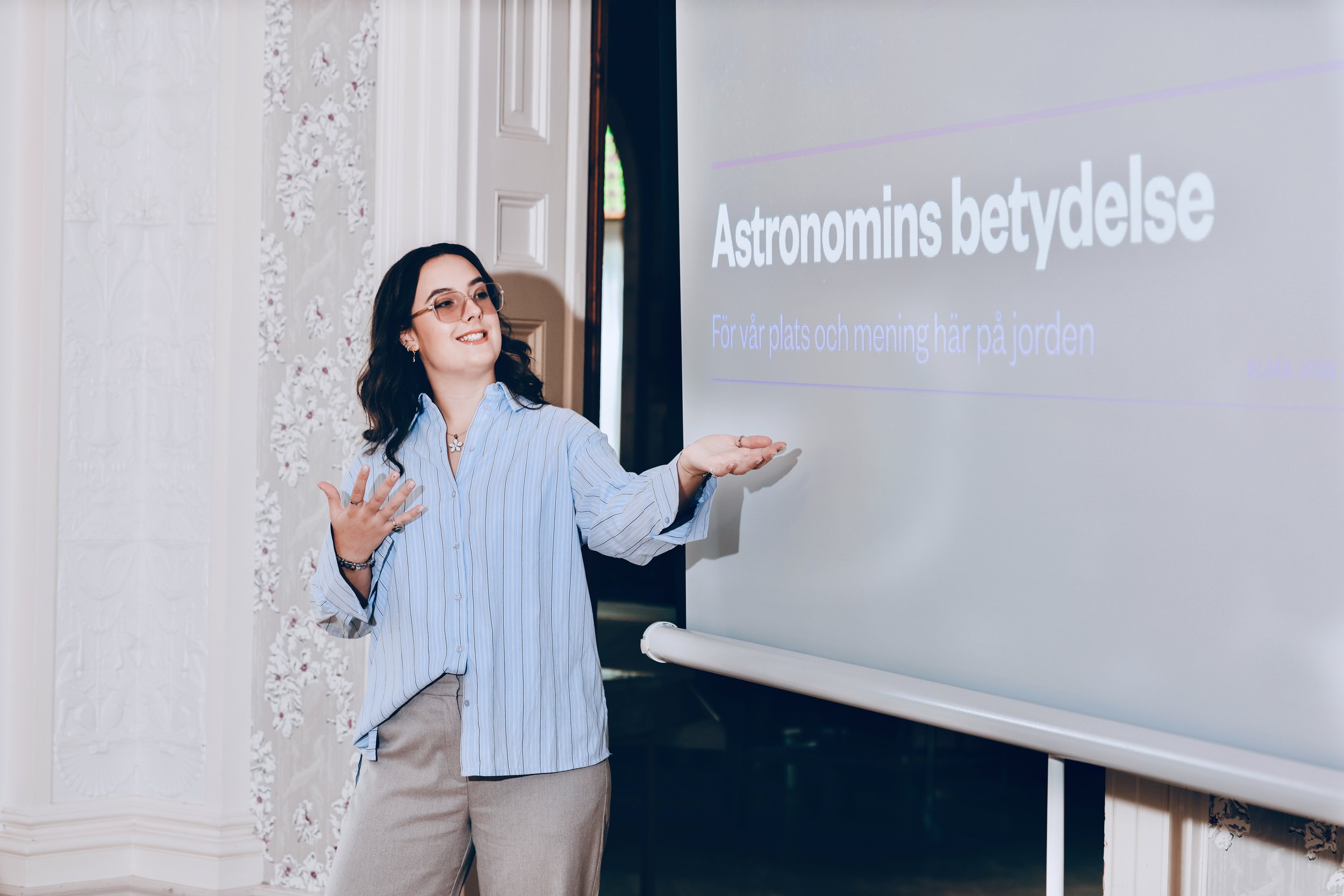 En kvinna håller ut handen som om hon presenterar en presentation framför en vägg med text om astronomi.