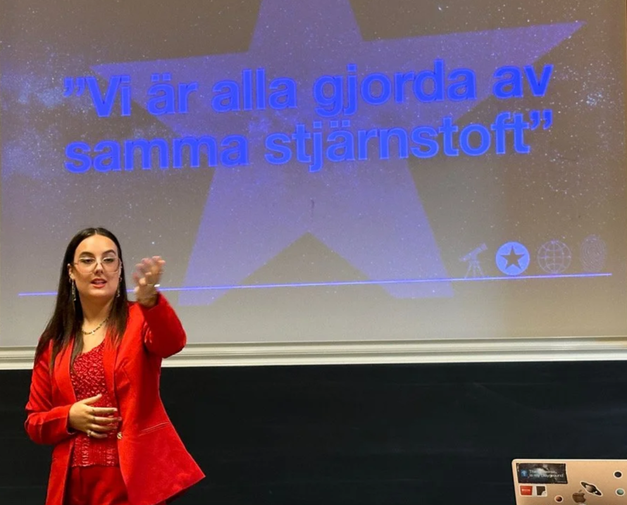 En kvinna i röd kappa som håller fram en hand framför en presentation som har texten 'Vi är alla gjorda av samma stjärnstoft' på en stjärnprydd bakgrund.