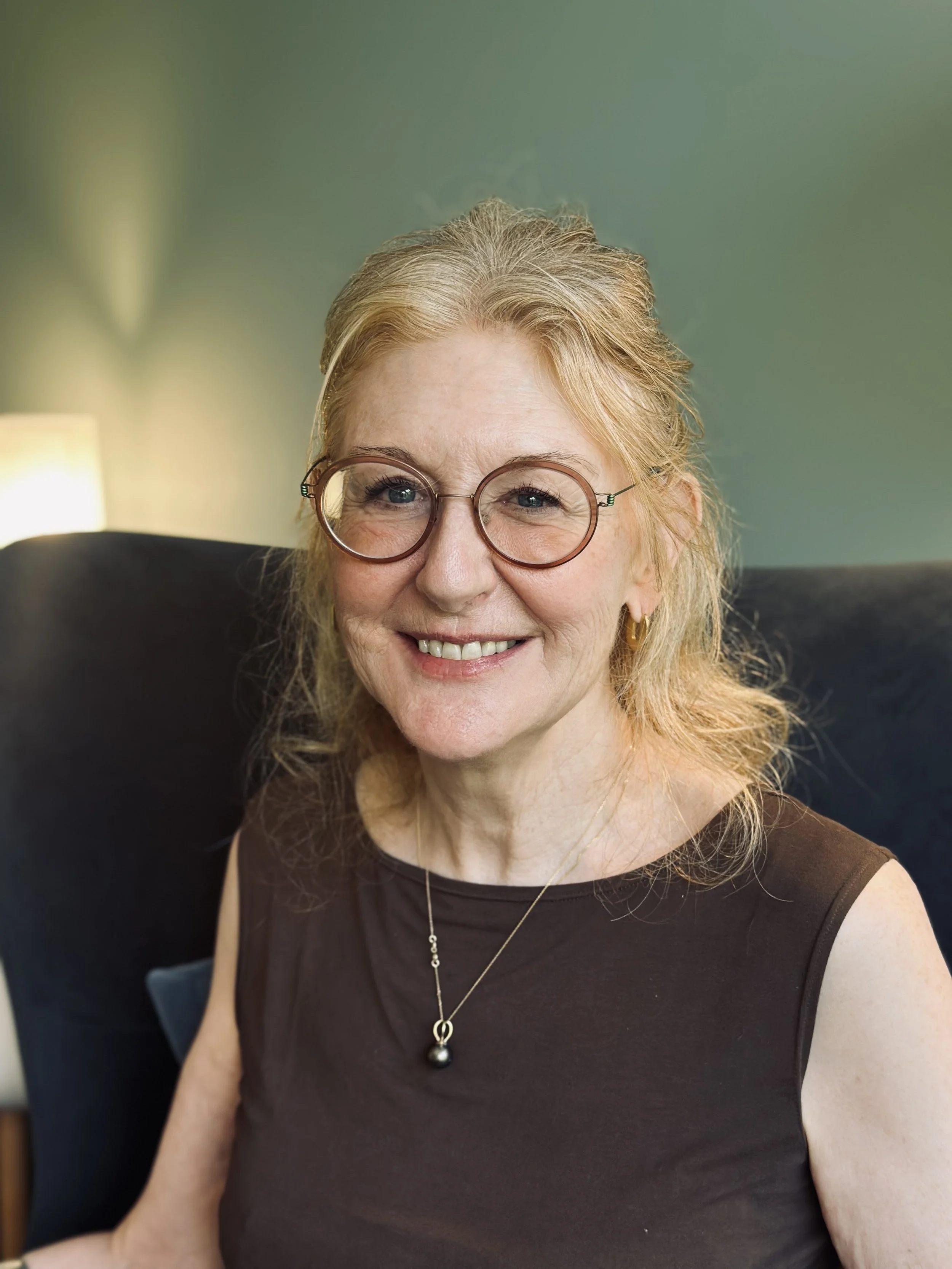 A woman with glasses, blonde hair, and a warm smile, wearing a dark sleeveless top, gold earrings, and a pendant necklace, sitting in a cozy room with a lamp and a green wall.