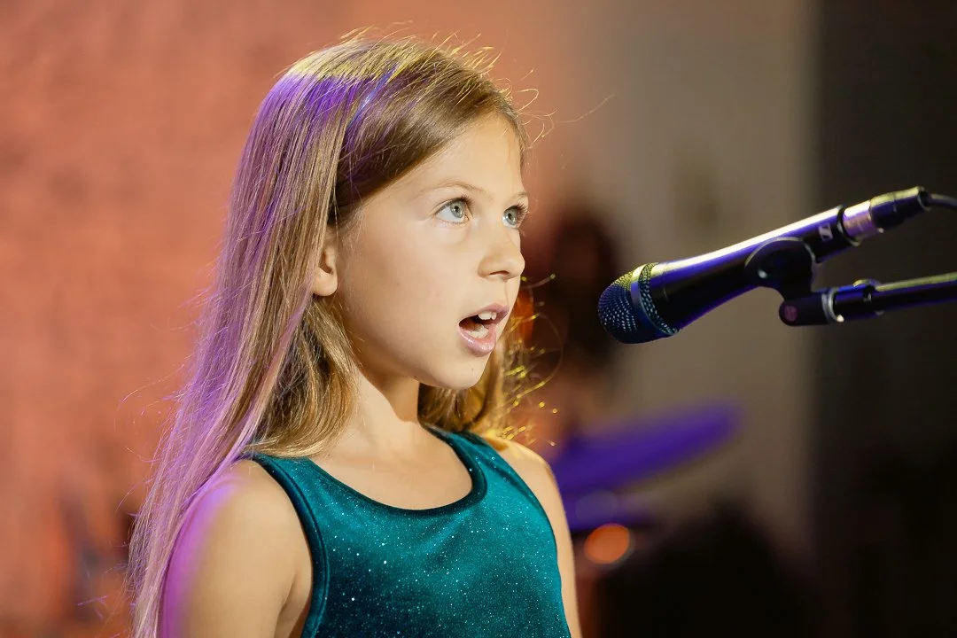 A young student from Noteworthy Music Academy in West Bloomfield singing into a microphone on stage.