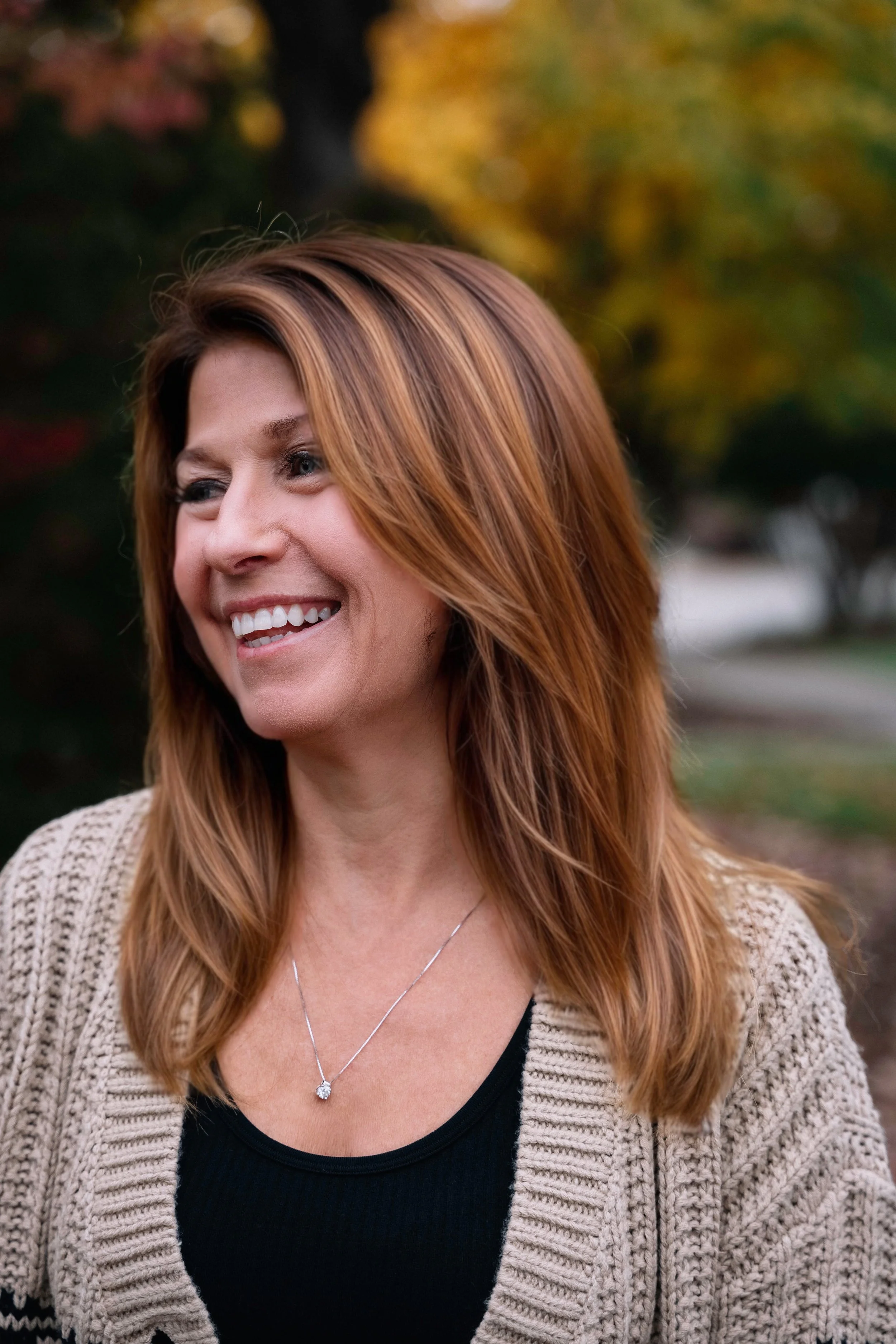 A woman with shoulder-length brown hair smiling outdoors during autumn, wearing a beige knitted cardigan, a black top, and a silver necklace with a diamond pendant.