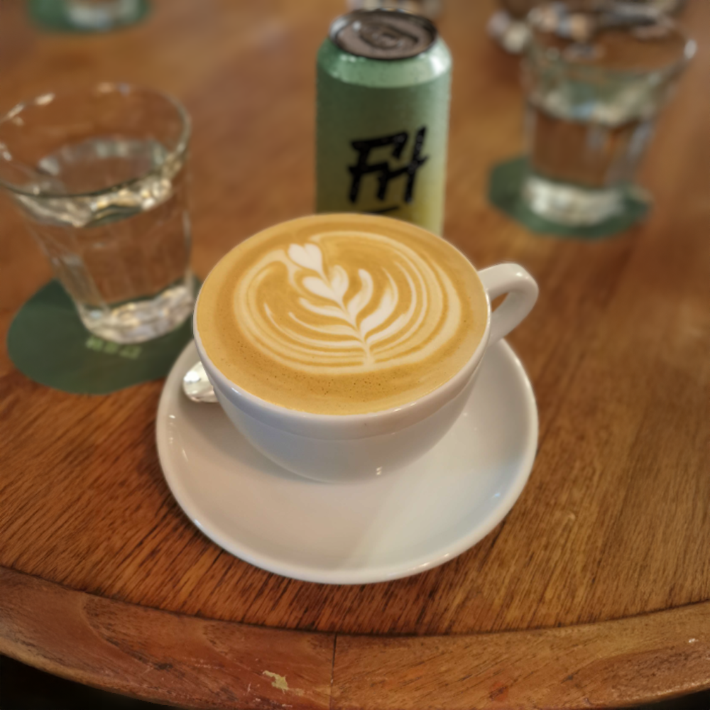 Un cappuccino avec un dessin sur la mousse, entouré de deux verres d'eau, une canette portant le logo 'FH', sur une table en bois.