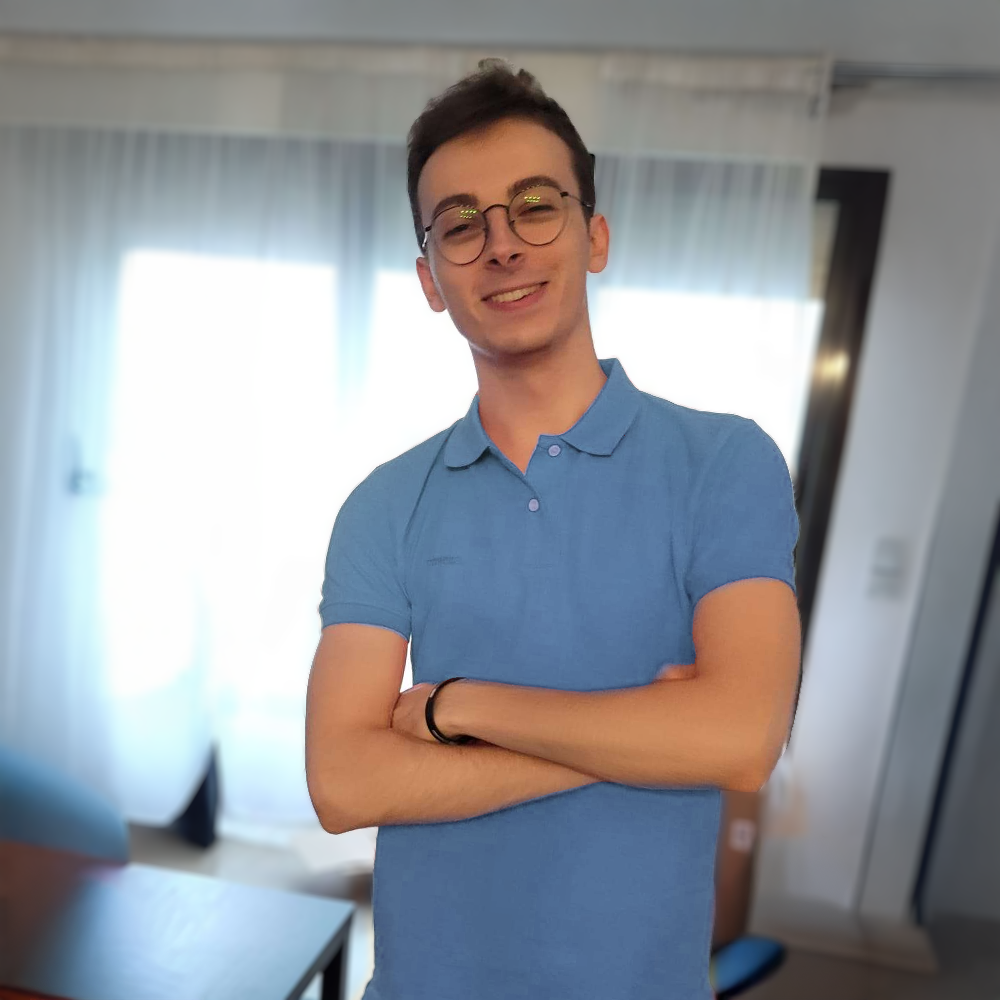 Un jeune homme souriant portant des lunettes, habillé d'un polo bleu, se tient dans une pièce lumineuse avec des rideaux blancs en arrière-plan.