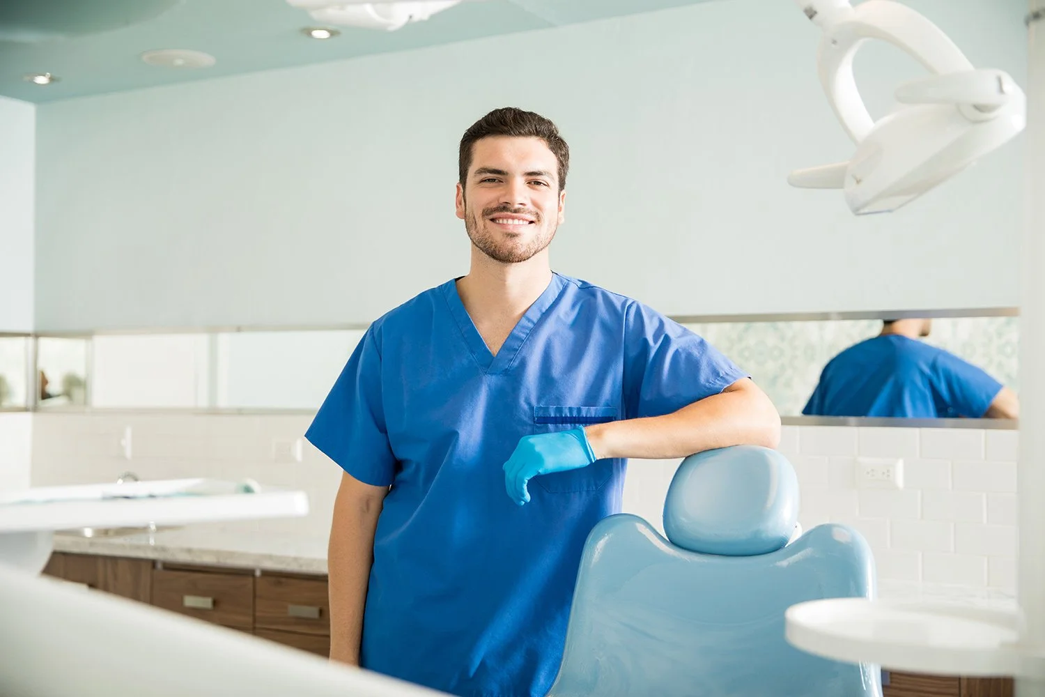 A smiling male dental professional standing in a dental clinic, wearing blue scrubs and a glove, with dental equipment and a dental chair in the background.