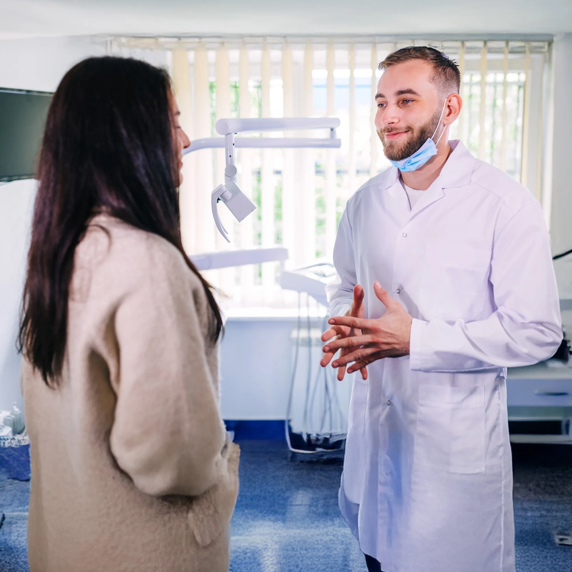A male healthcare professional in a white coat and face mask talking to a female patient in a beige coat, inside a medical office with dental equipment in the background.
