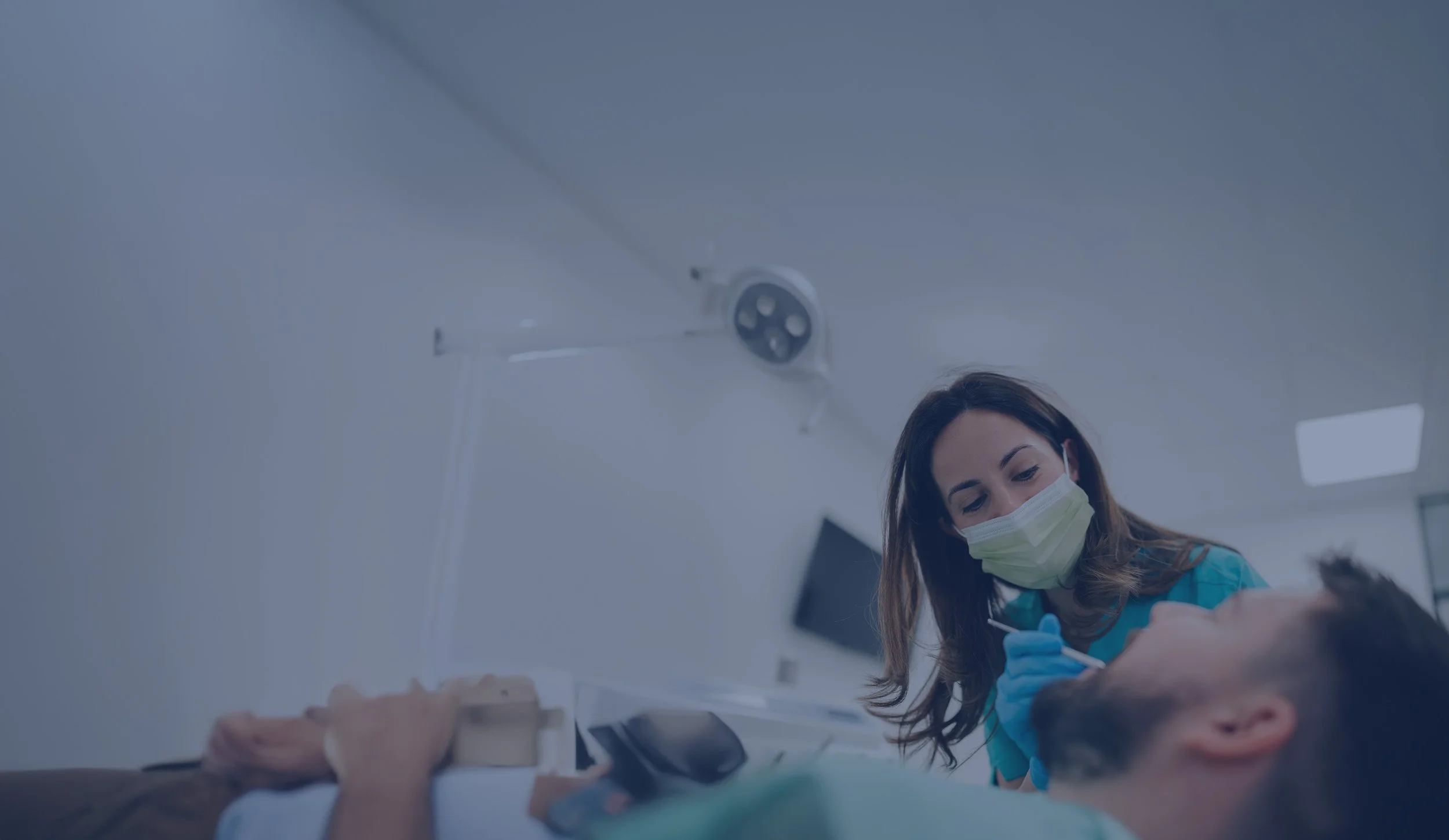 A female nurse wearing a medical mask and gloves administering an injection to a man lying in a hospital bed.