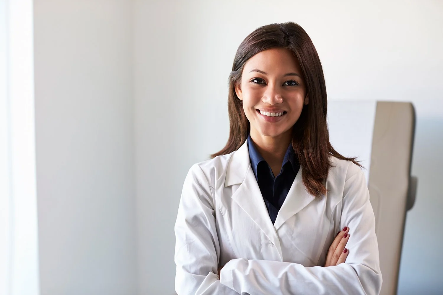 A young woman healthcare professional smiling, wearing a white coat and dark shirt, in an office setting.